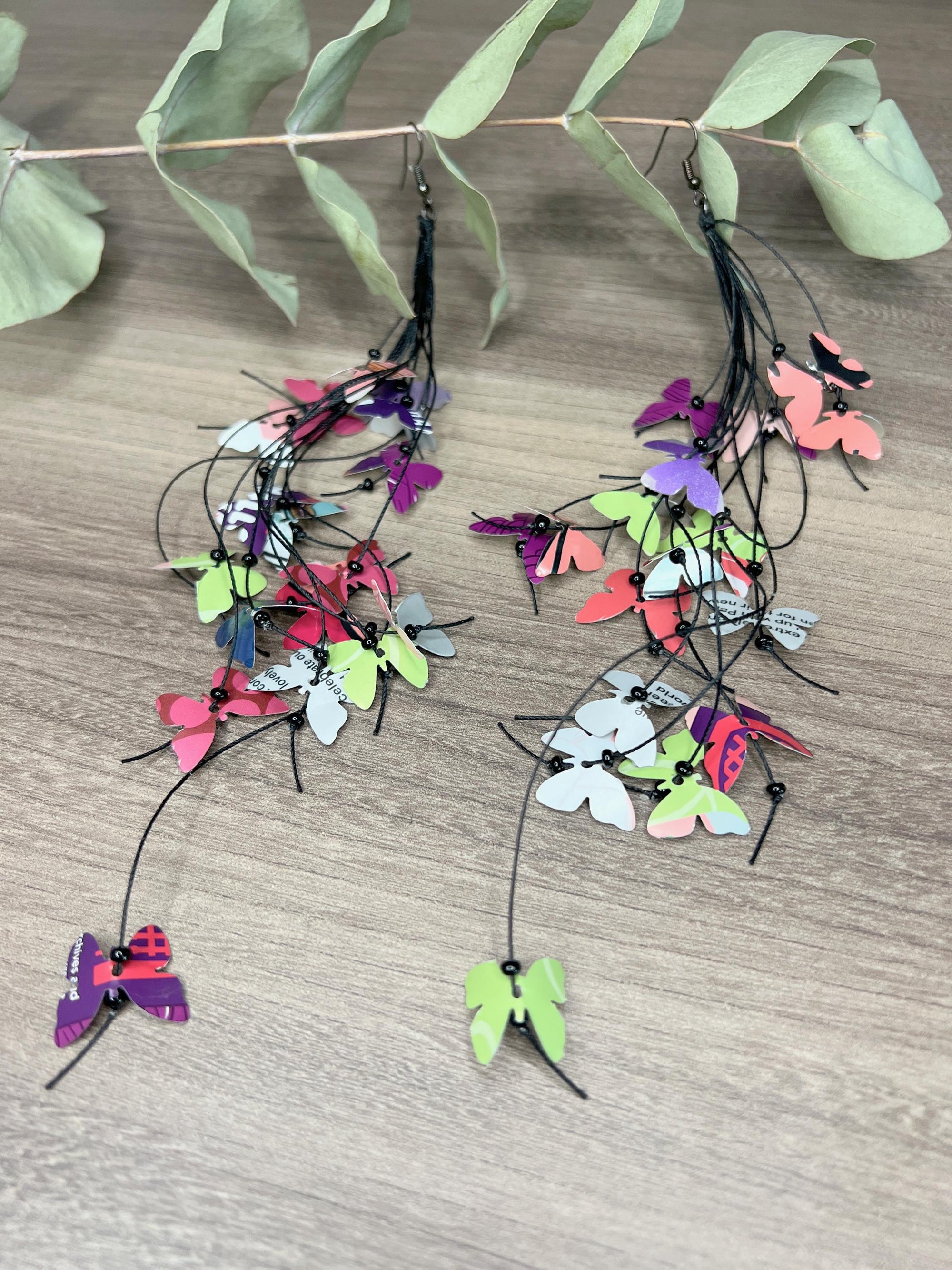extra long, upcycled earrings hanging on a dry wooden stick with leaves, the earrings feature tiny butterflies cut from soda and beer cans, fitted on waxed cotton cord