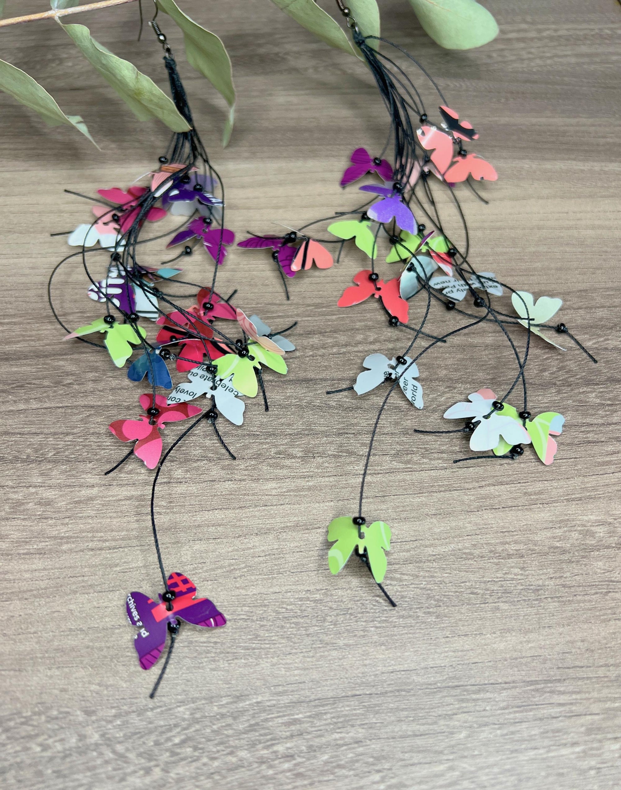 extra long, upcycled earrings hanging on a dry wooden stick with leaves, the earrings feature tiny butterflies cut from soda and beer cans, fitted on waxed cotton cord