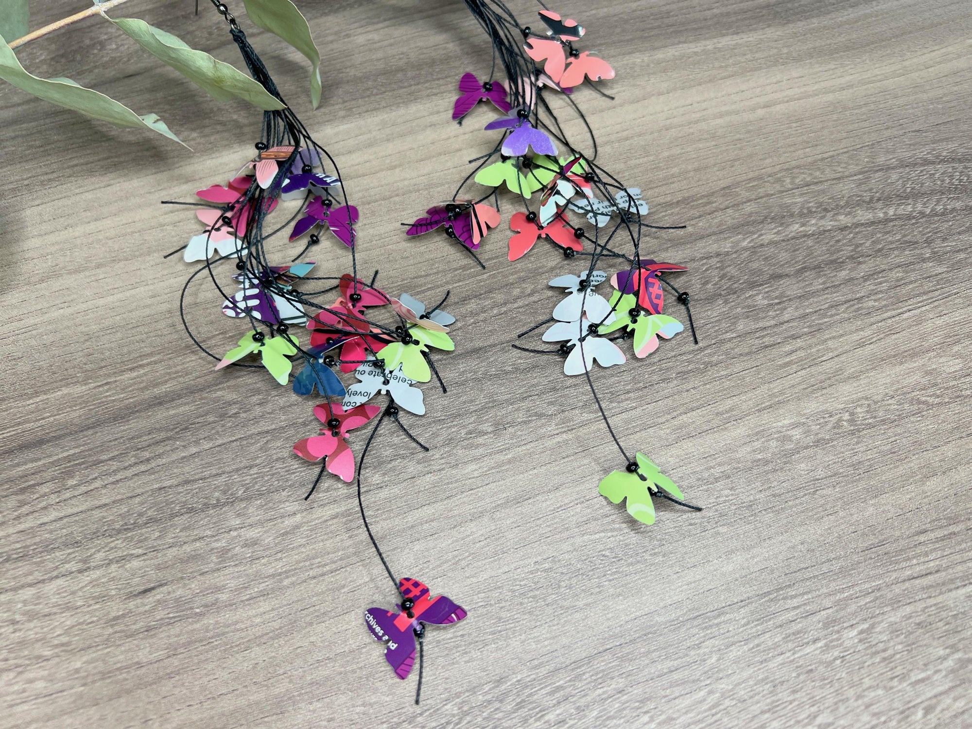 extra long, upcycled earrings hanging on a dry wooden stick with leaves, the earrings feature tiny butterflies cut from soda and beer cans, fitted on waxed cotton cord