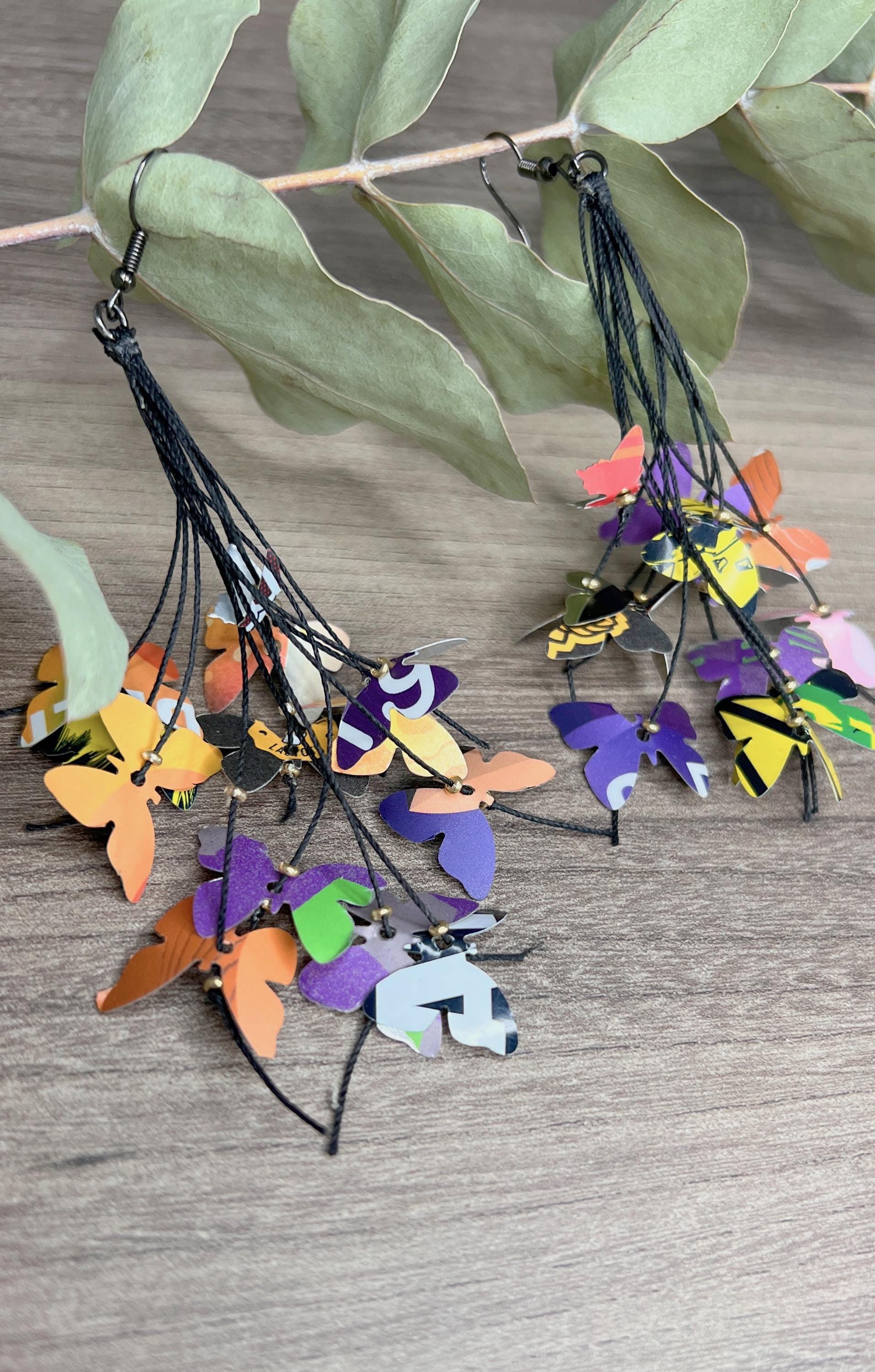 Upcycled earrings hanging on a dry wooden stick with leaves, the earrings feature tiny butterflies cut from soda and beer cans, fitted on waxed cotton cord