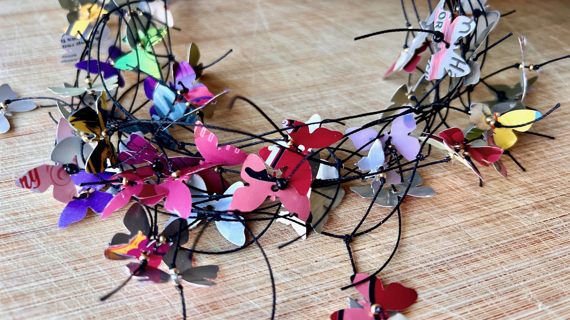 elegant, colourful necklace featuring tiny aluminium butterflies, each one is cut from soda and beer cans, fitted on multiple thin black waxed cotton cord on a wooden background