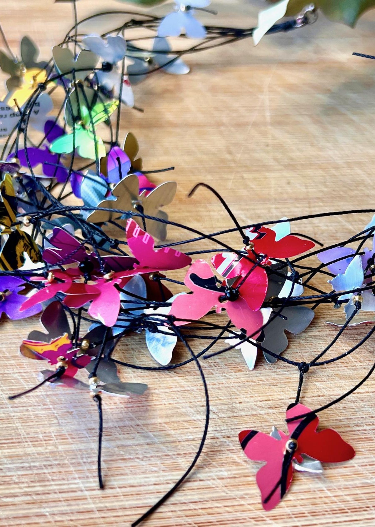 elegant, colourful necklace featuring tiny aluminium butterflies, each one is cut from soda and beer cans, fitted on multiple thin black waxed cotton cord on a wooden background