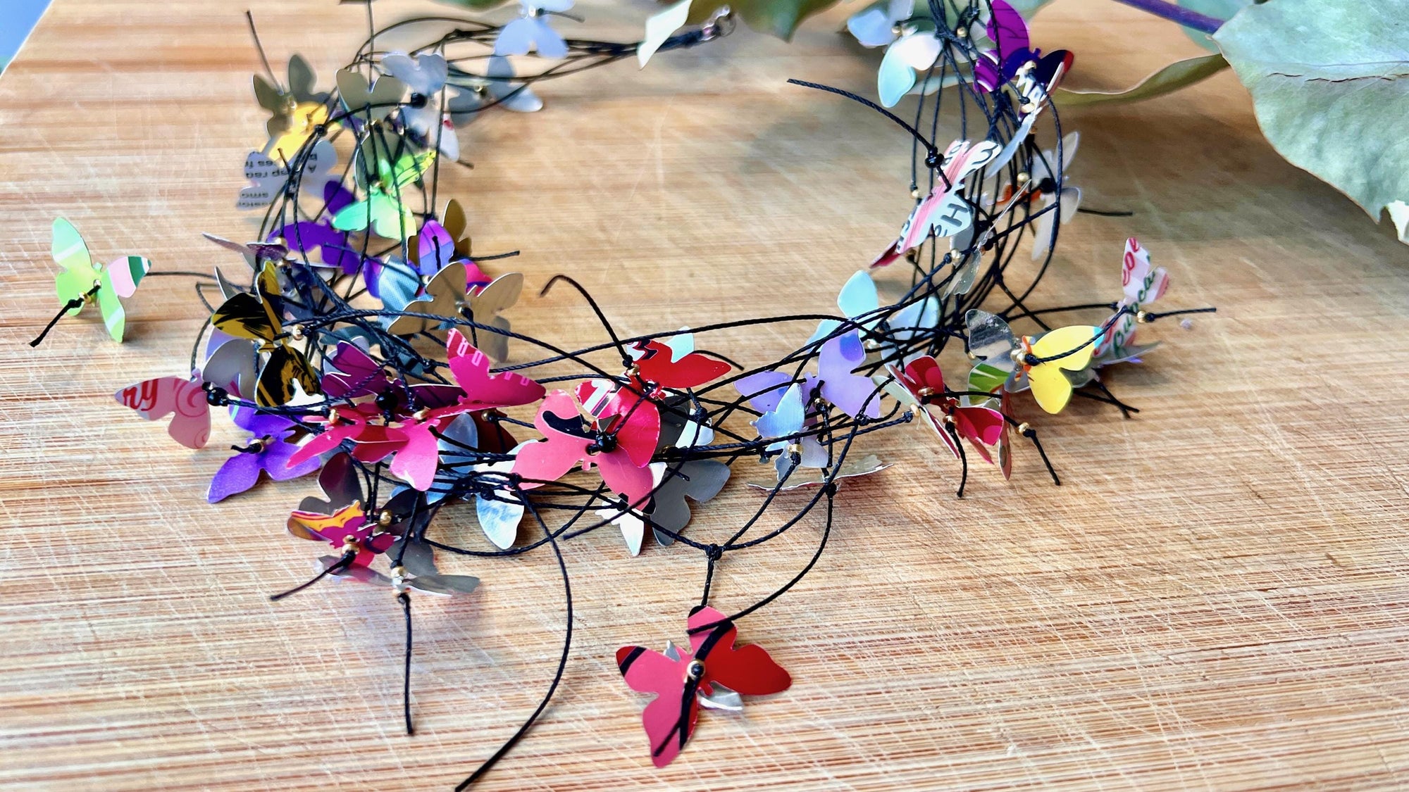 elegant, colourful necklace featuring tiny aluminium butterflies, each one is cut from soda and beer cans, fitted on multiple thin black waxed cotton cord on a wooden background