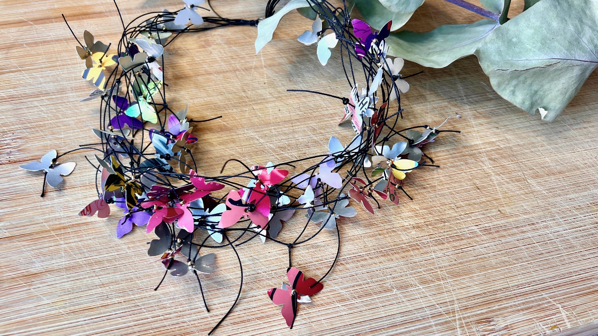 elegant, colourful necklace featuring tiny aluminium butterflies, each one is cut from soda and beer cans, fitted on multiple thin black waxed cotton cord on a wooden background