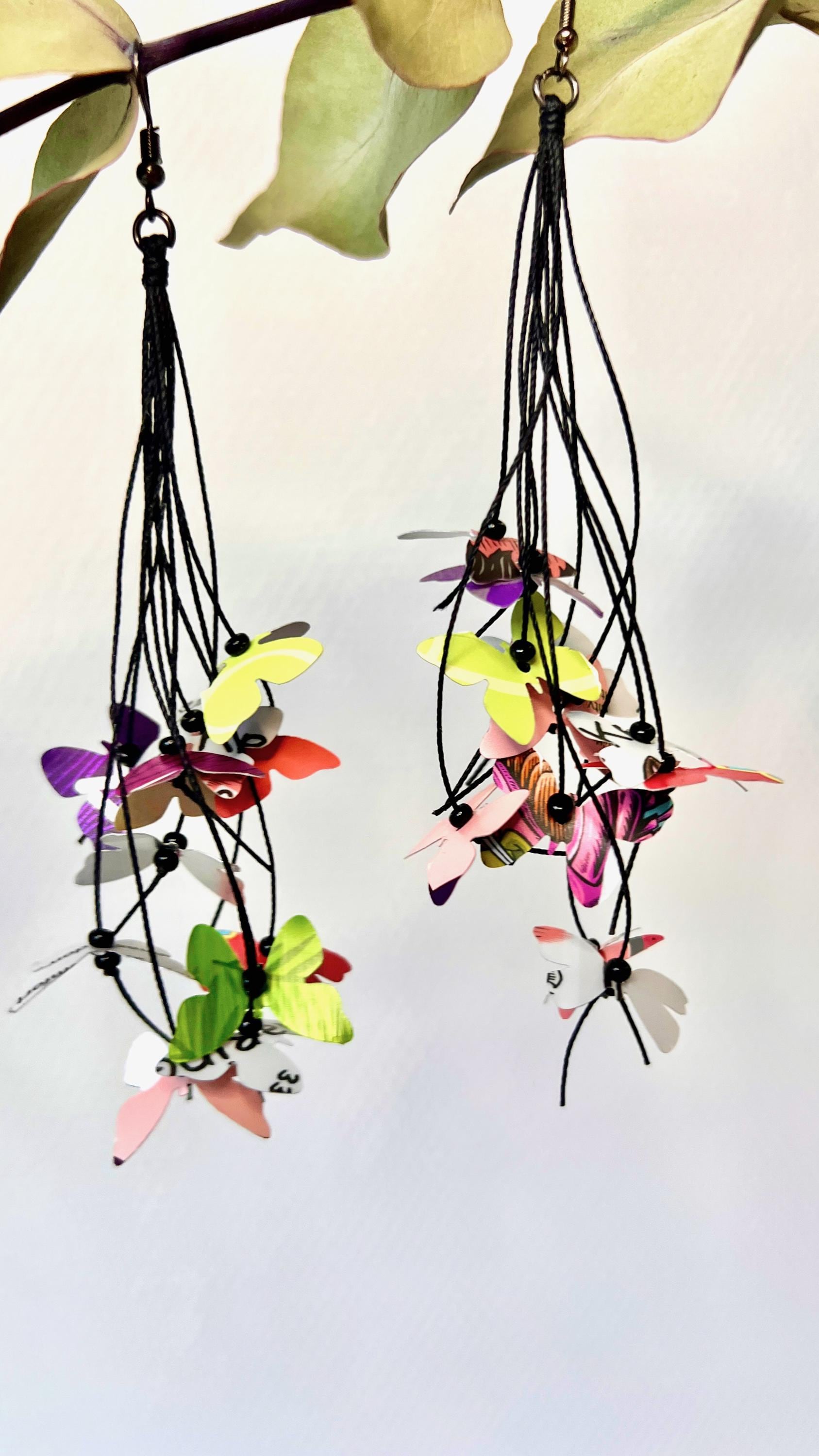 Upcycled earrings hanging on a dry wooden stick with leaves, the earrings feature tiny butterflies cut from soda and beer cans, fitted on waxed cotton cord