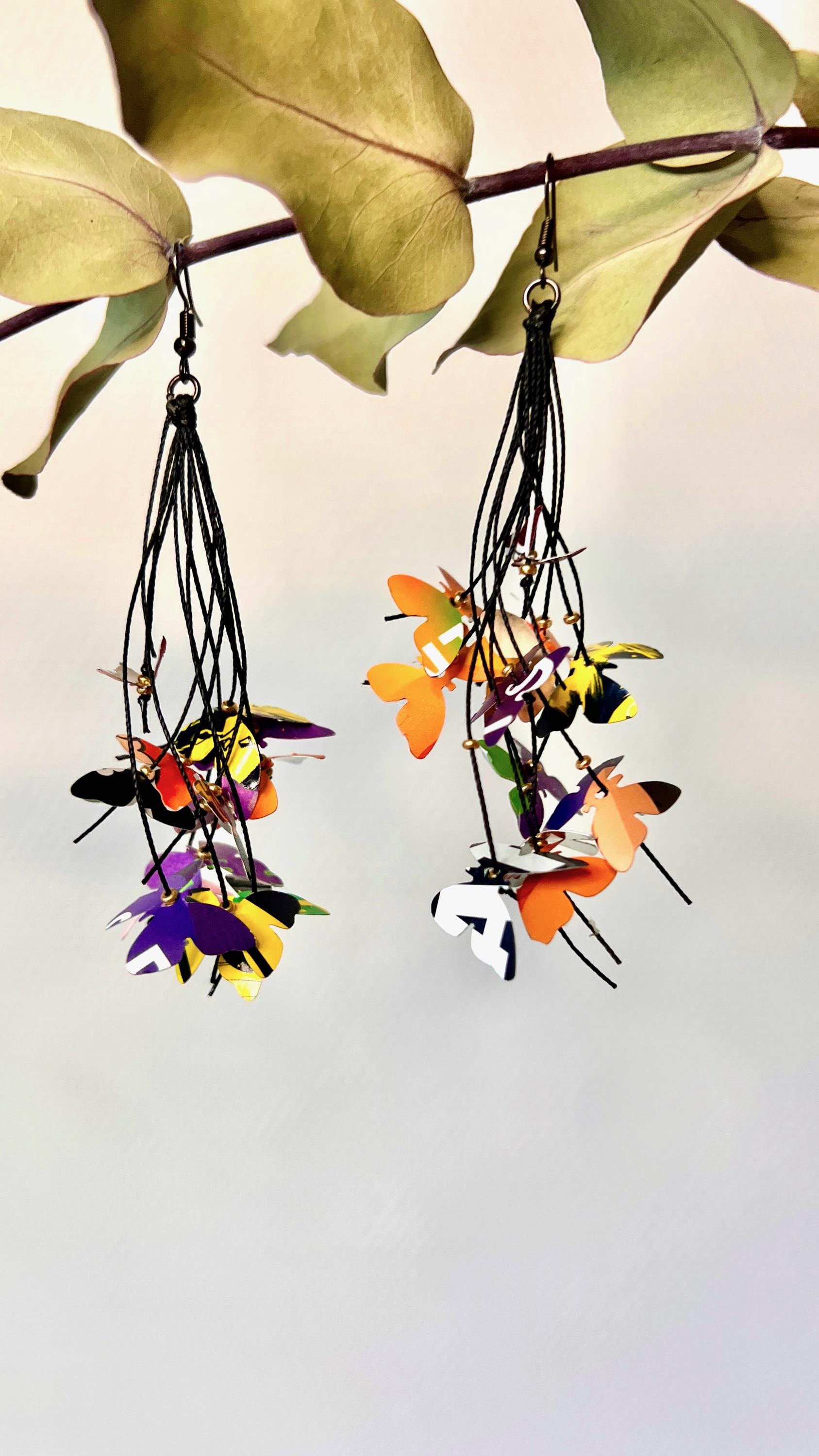 Upcycled earrings hanging on a dry wooden stick with leaves, the earrings feature tiny butterflies cut from soda and beer cans, fitted on waxed cotton cord