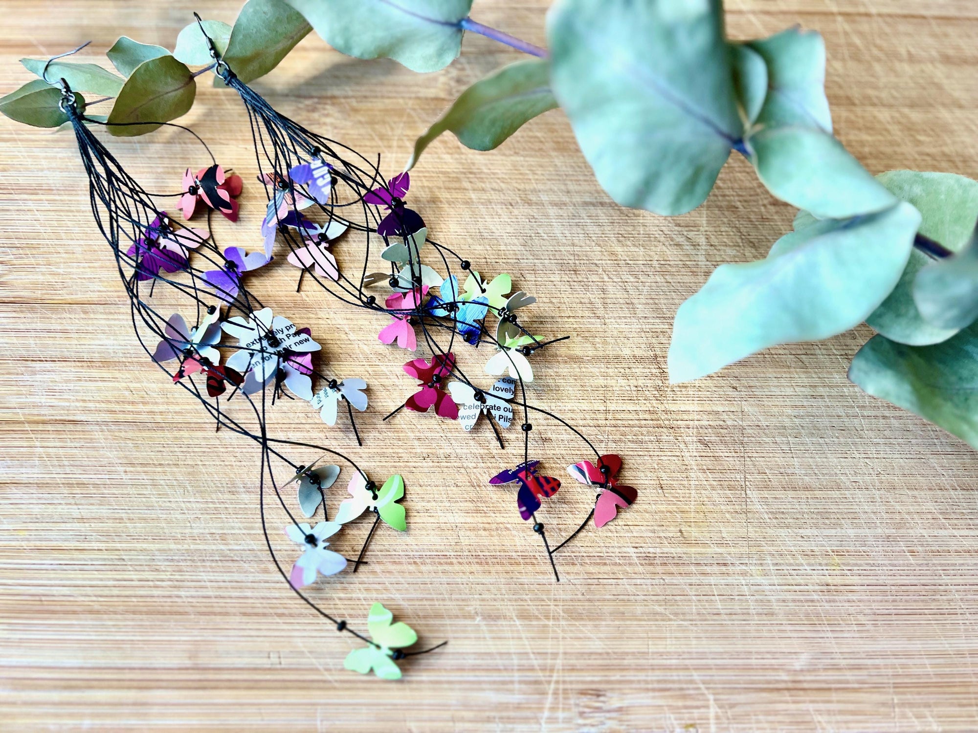 Extra long, Upcycled earrings on a wooden backround , the earrings feature tiny butterflies cut from soda and beer cans, fitted on waxed cotton cord