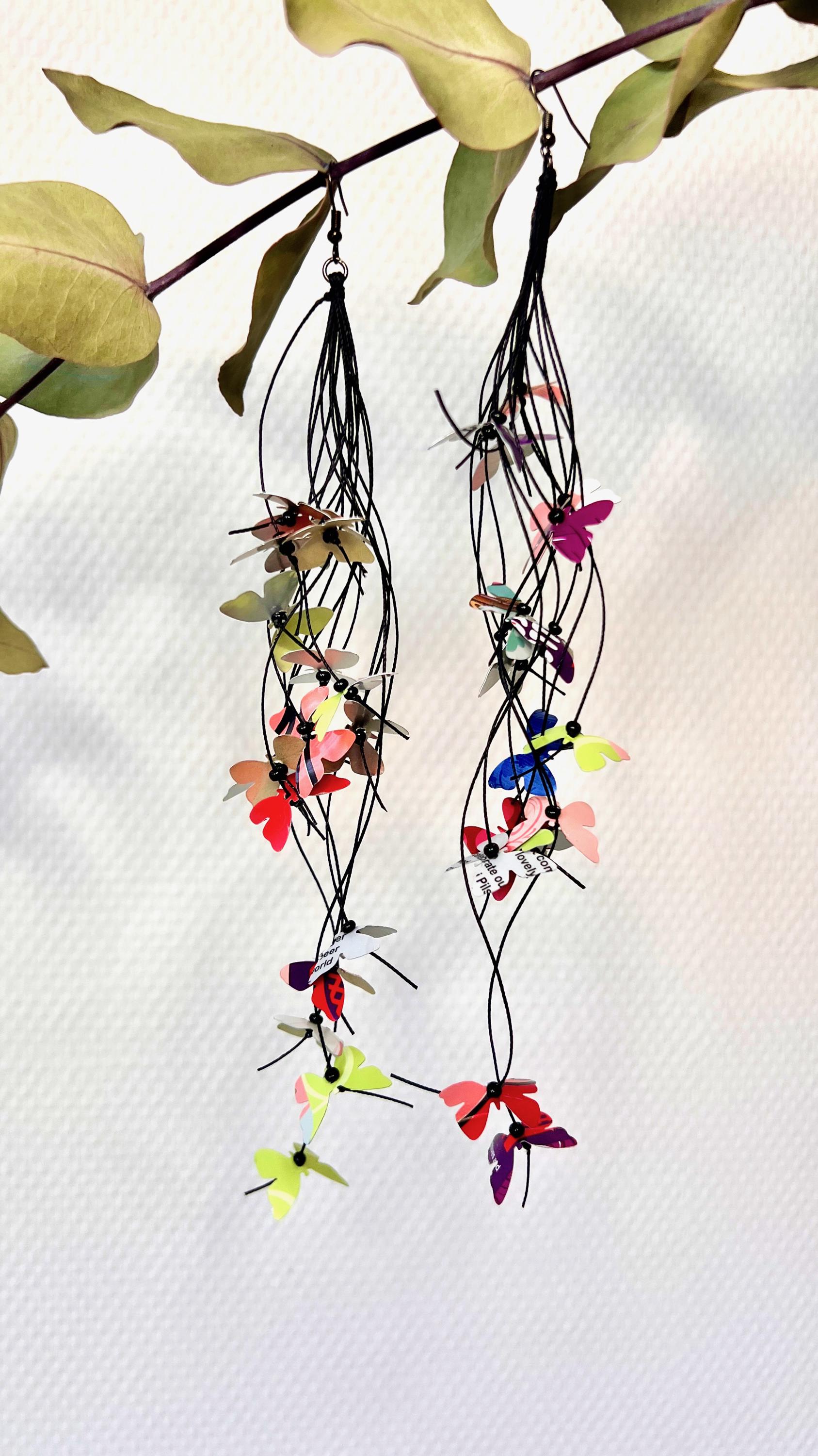 extra long, upcycled earrings hanging on a dry wooden stick with leaves, the earrings feature tiny butterflies cut from soda and beer cans, fitted on waxed cotton cord