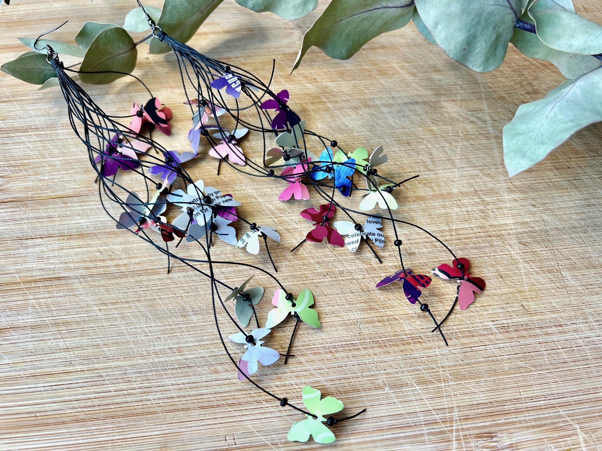 Extra long, Upcycled earrings on a wooden backround , the earrings feature tiny butterflies cut from soda and beer cans, fitted on waxed cotton cord