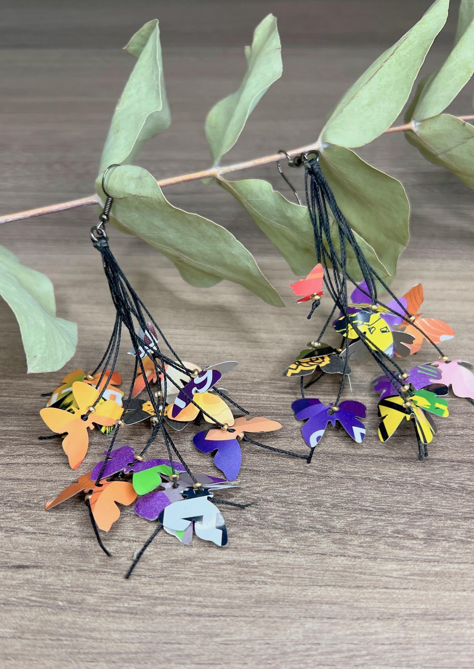 Upcycled earrings hanging on a dry wooden stick with leaves, the earrings feature tiny butterflies cut from soda and beer cans, fitted on waxed cotton cord