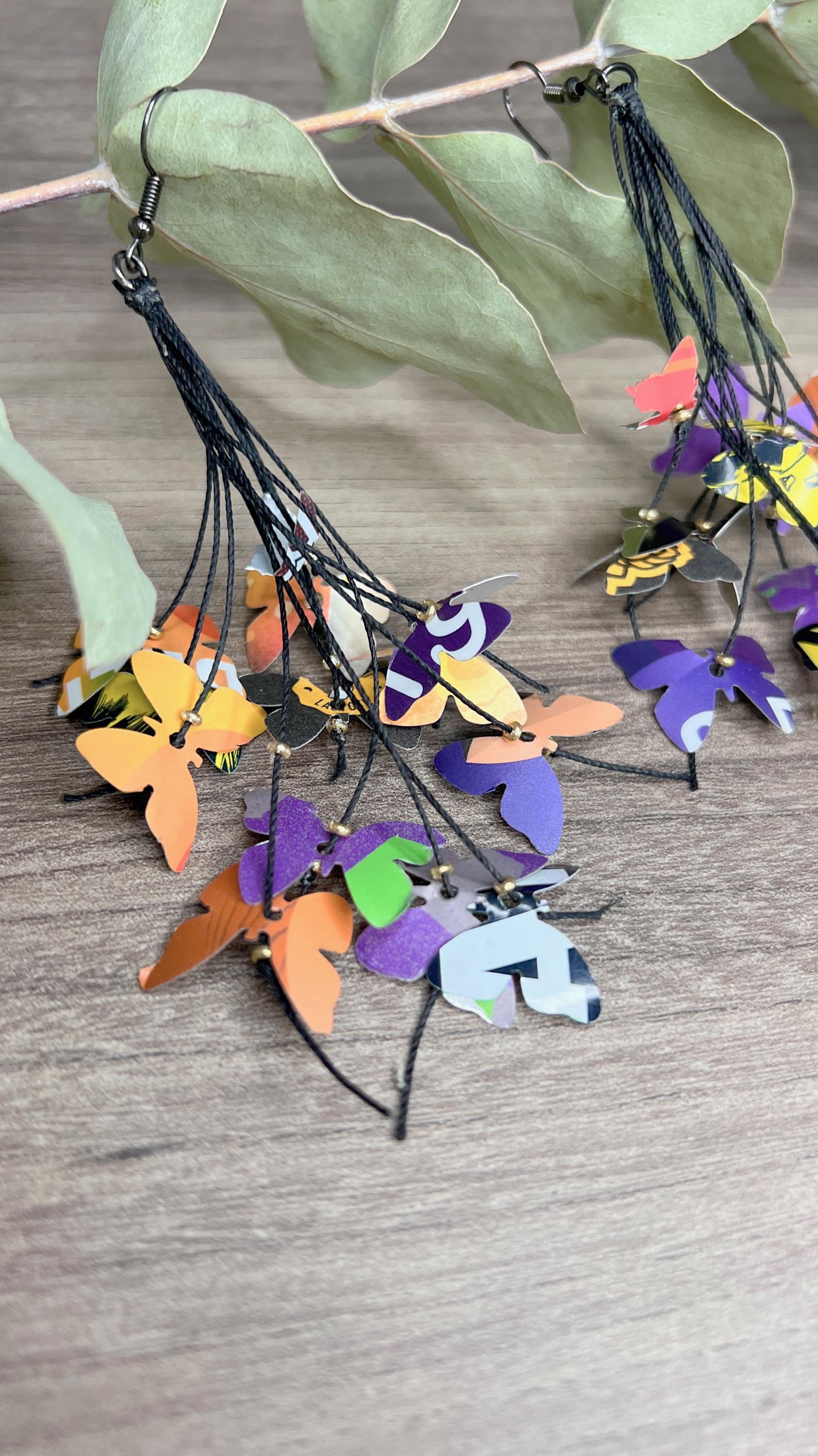 Upcycled earrings hanging on a dry wooden stick with leaves, the earrings feature tiny butterflies cut from soda and beer cans, fitted on waxed cotton cord