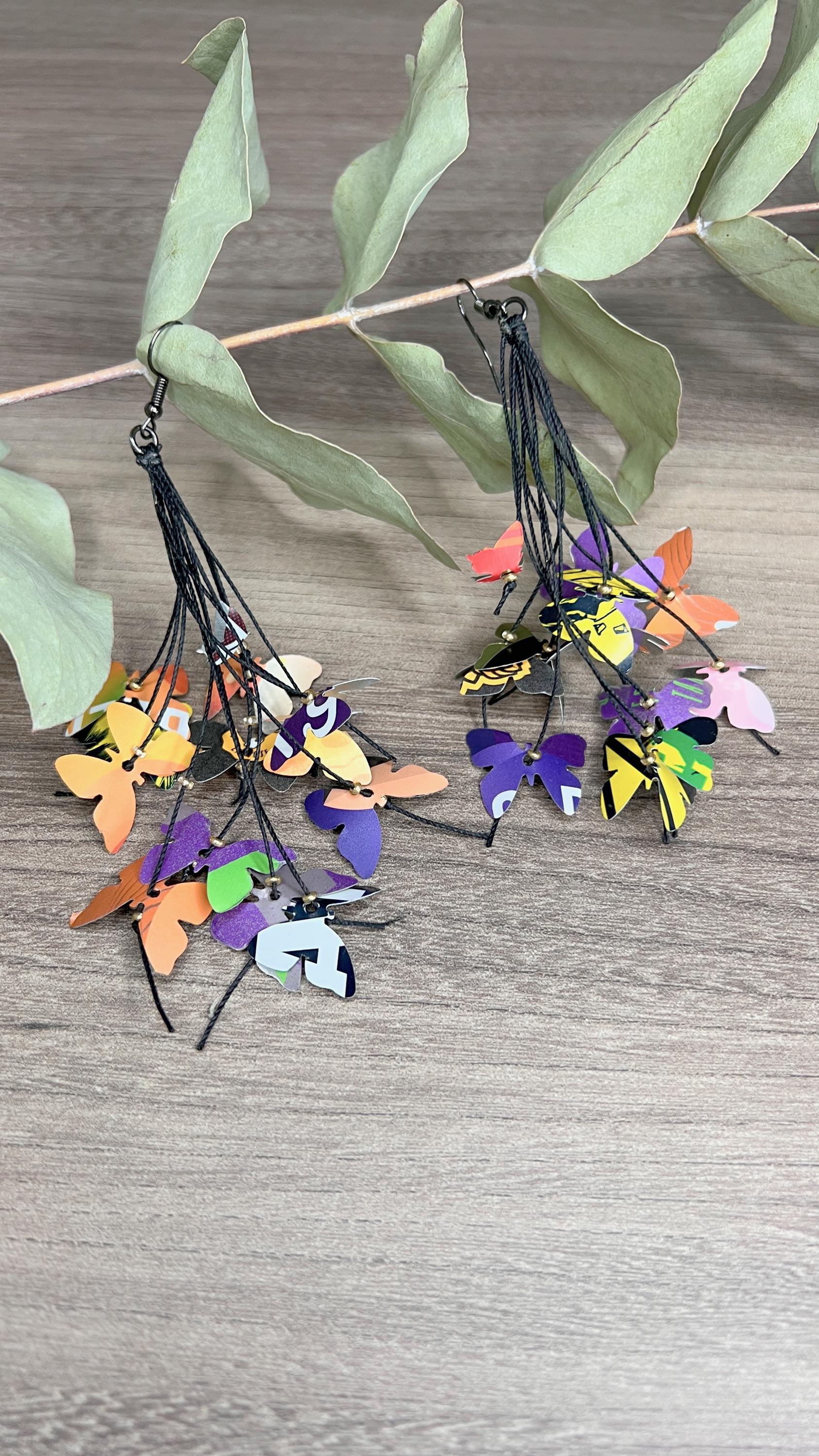 Upcycled earrings hanging on a dry wooden stick with leaves, the earrings feature tiny butterflies cut from soda and beer cans, fitted on waxed cotton cord