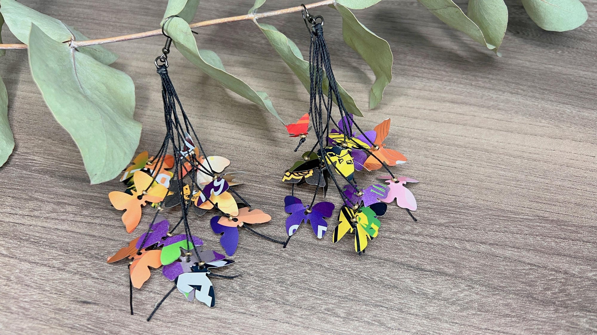Upcycled earrings hanging on a dry wooden stick with leaves, the earrings feature tiny butterflies cut from soda and beer cans, fitted on waxed cotton cord