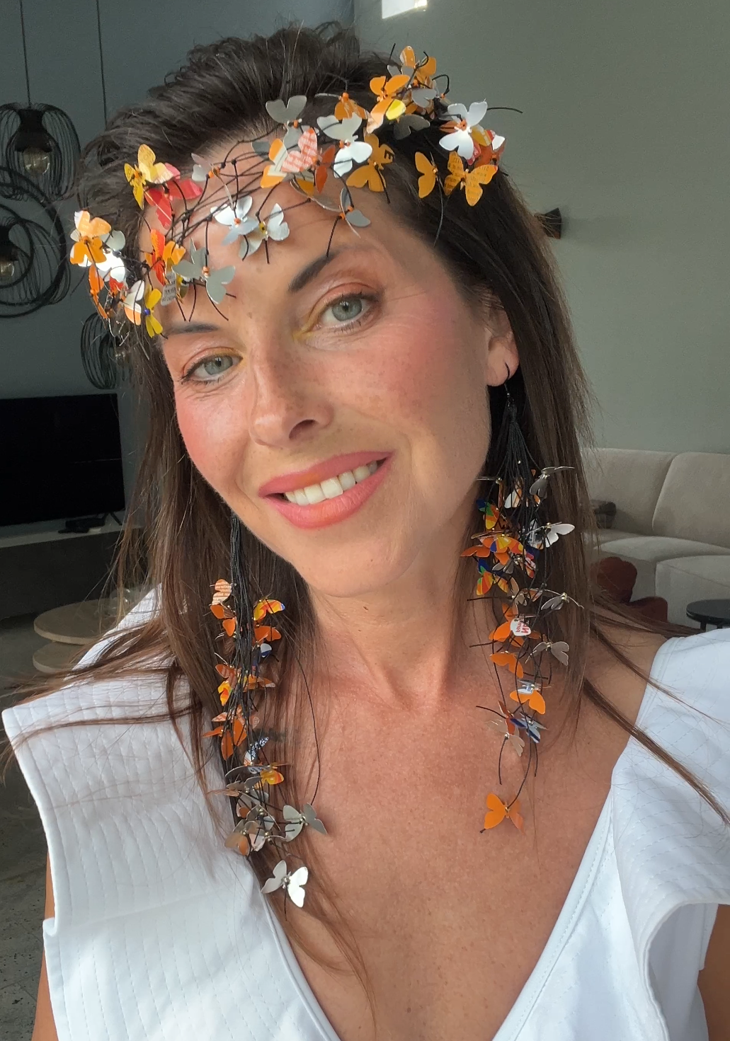 A woman wearing an upcycled, decorative butterfly crown and matching butterfly necklace, made from fanta cans  photographed in a real indoor setting with a window and wall visible in the background.