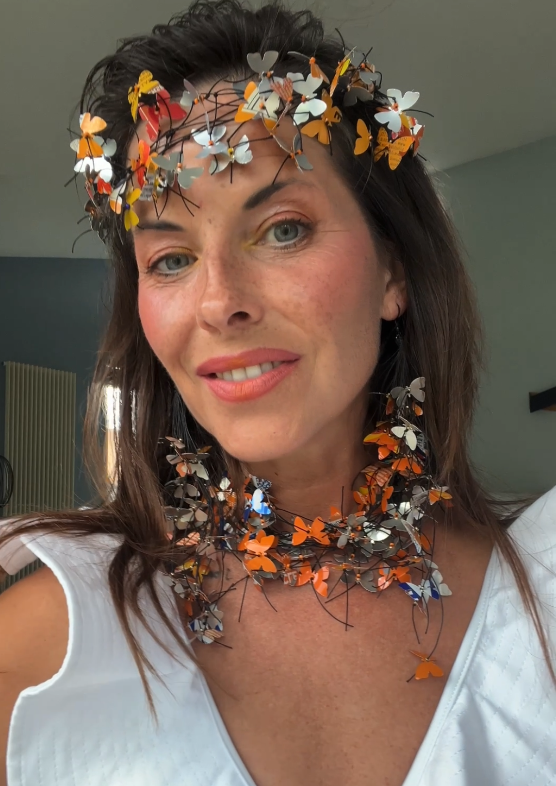 A woman wearing an upcycled, decorative butterfly crown and matching butterfly necklace, made from fanta cans  photographed in a real indoor setting with a window and wall visible in the background.