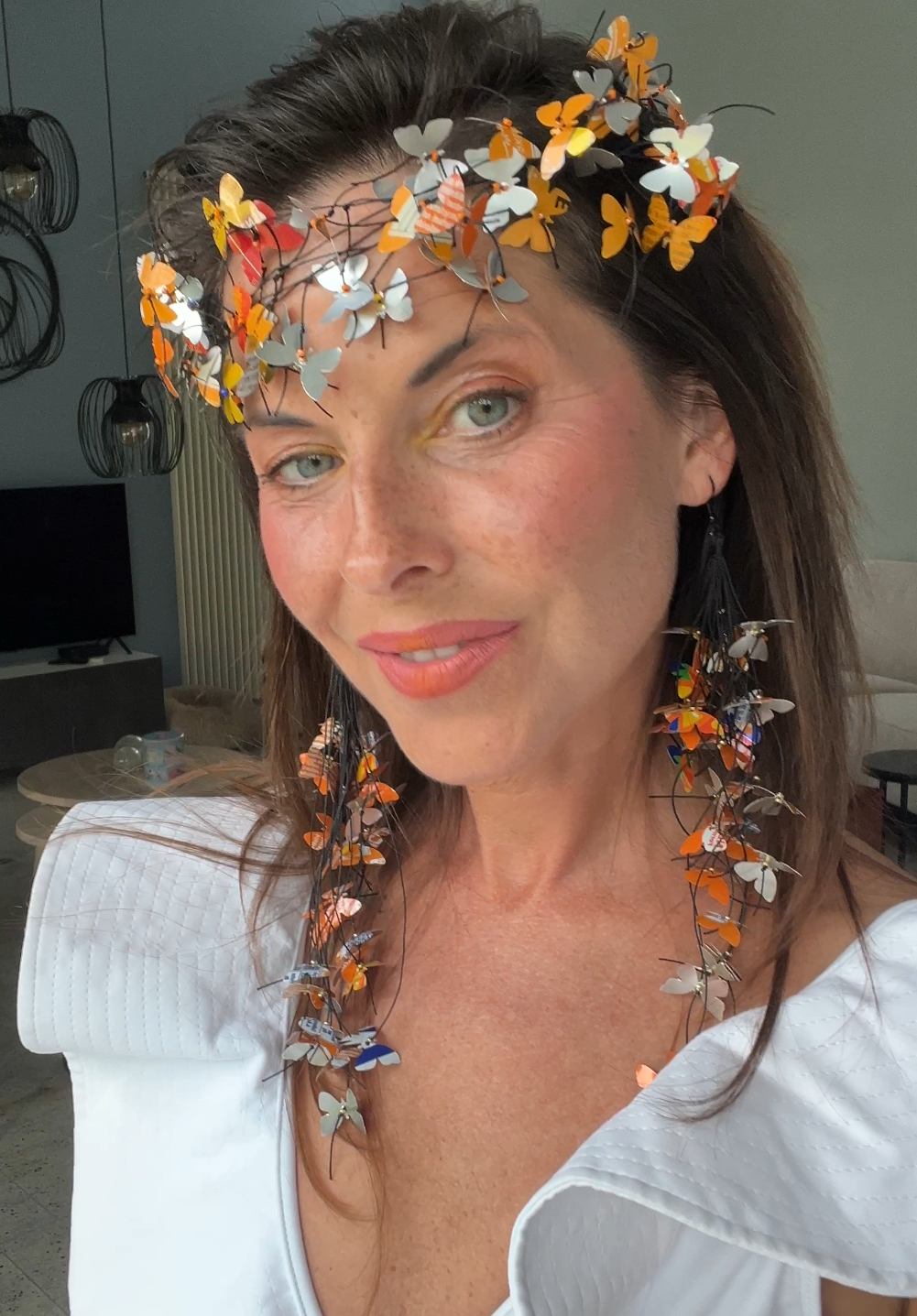 A woman wearing an upcycled, decorative butterfly crown and matching butterfly necklace, made from fanta cans  photographed in a real indoor setting with a window and wall visible in the background.