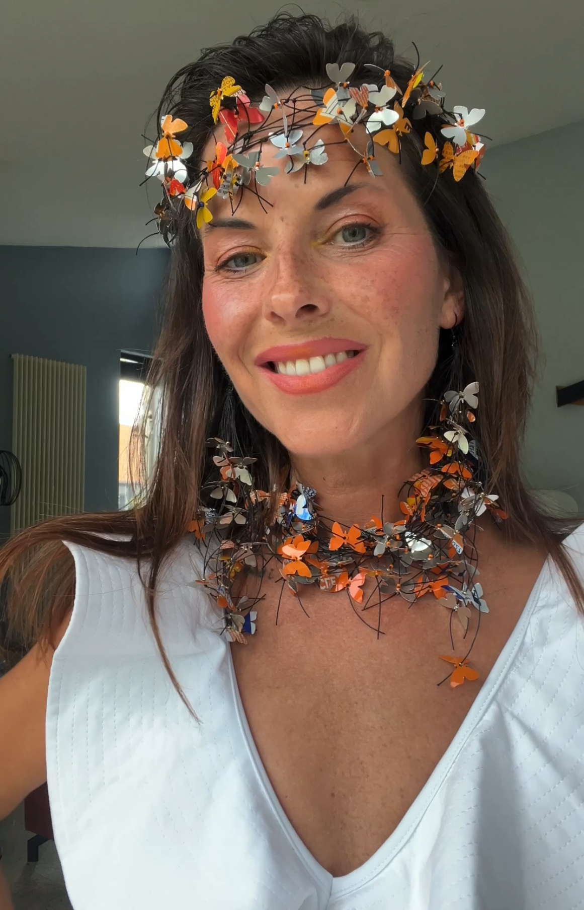A woman wearing an upcycled, decorative butterfly crown and matching butterfly necklace, made from fanta cans  photographed in a real indoor setting with a window and wall visible in the background.