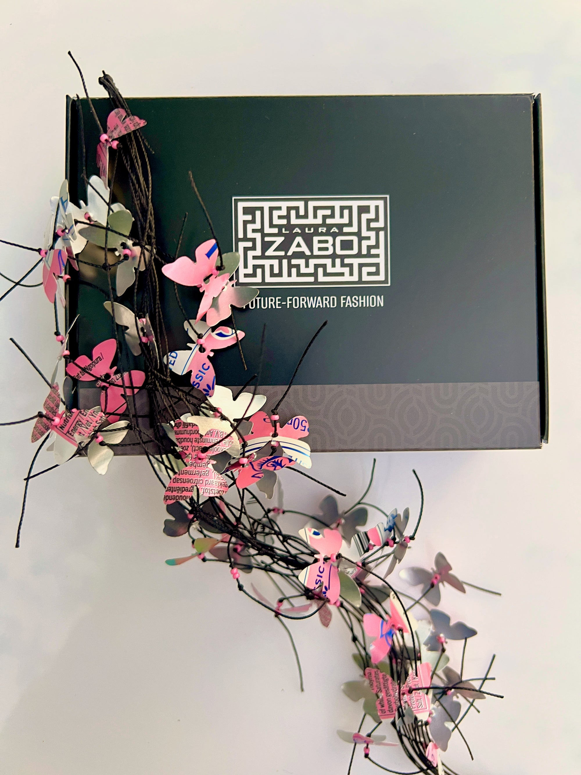 A black Zabo fashion box with a white geometric logo, decorated with pink, white, and gray butterfly upcycled necklace on a light gray background.