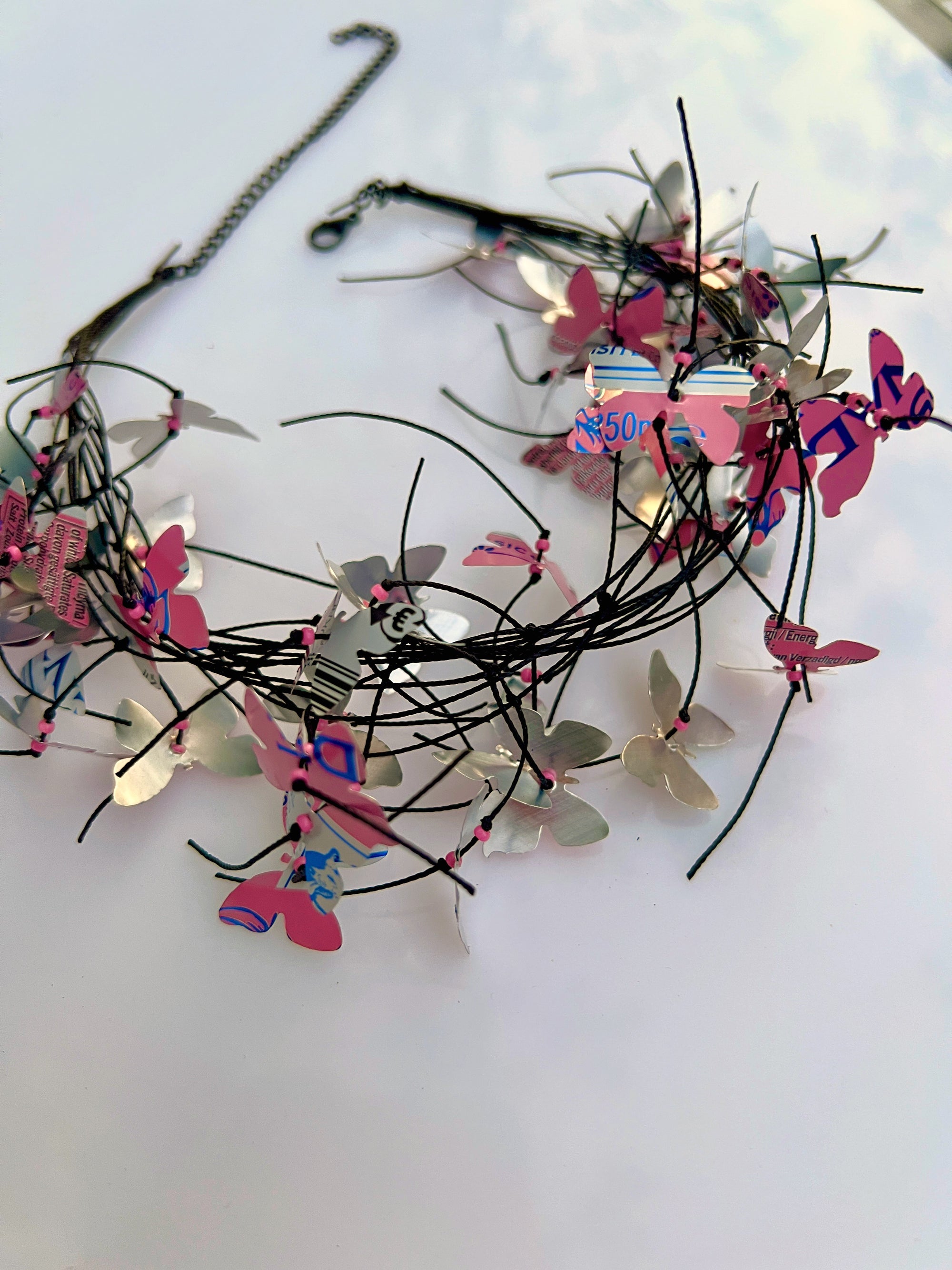 A handmade wire crown necklace decorated with metallic silver butterfly wings, pink paper butterflies, and small colorful printed pieces, displayed on a white surface.