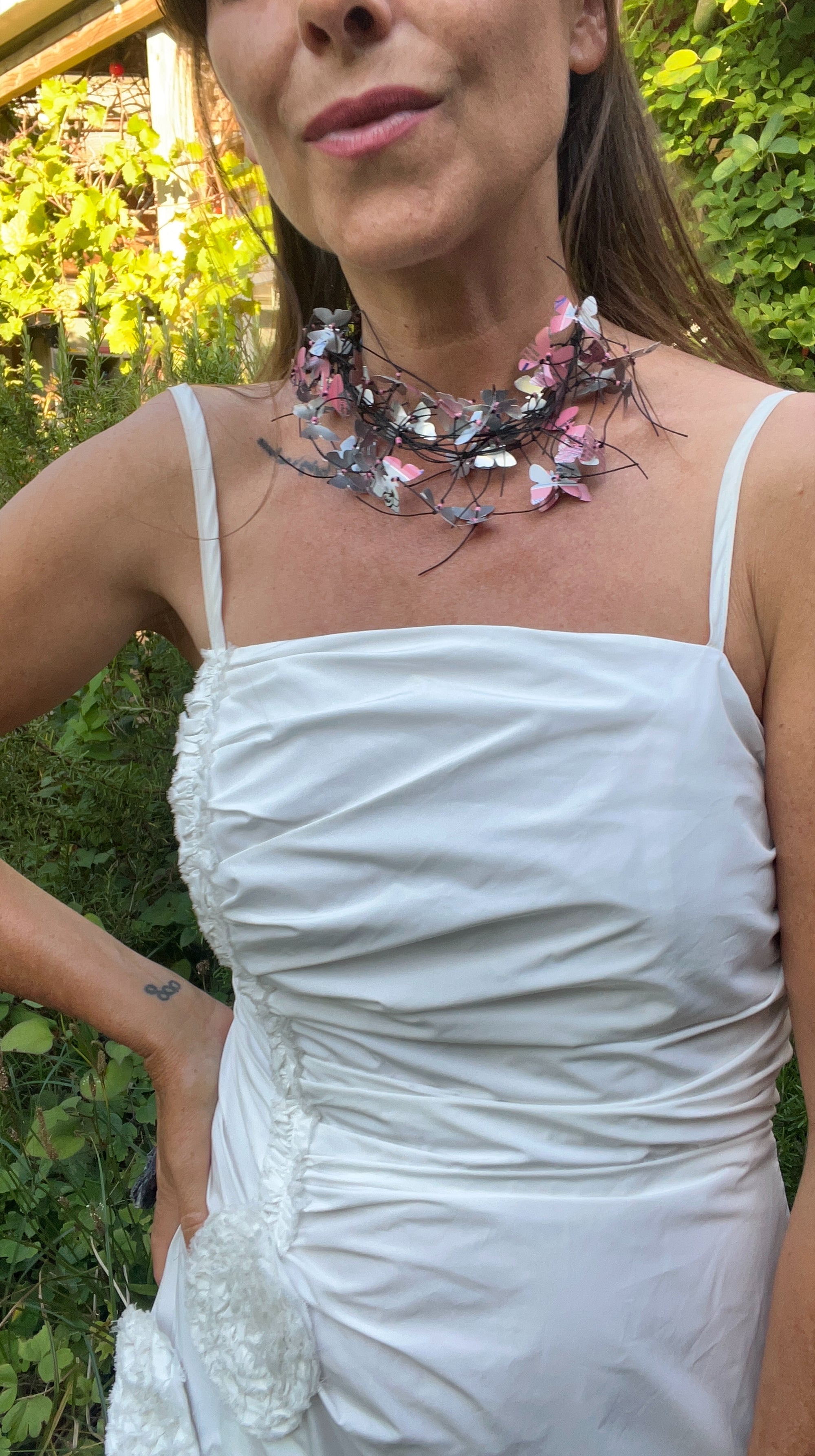 A woman wearing a white spaghetti-strap dress and a chunky multi-strand butterfly necklace made of small pink, blue, and gray butterflies cut from soda can on dark cotton cord, photographed outdoors in front of green foliage.