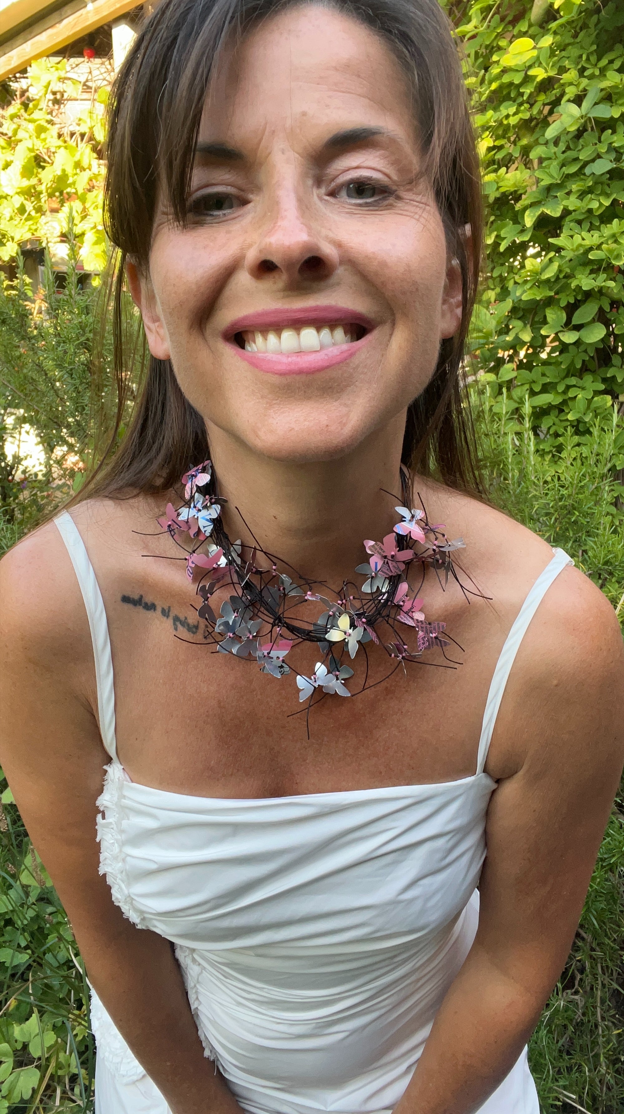 A smiling woman outdoors wearing a handmade butterfly collar necklace with pink butterflies cut from a soda can on a dark cord base, paired with a white spaghetti-strap top.