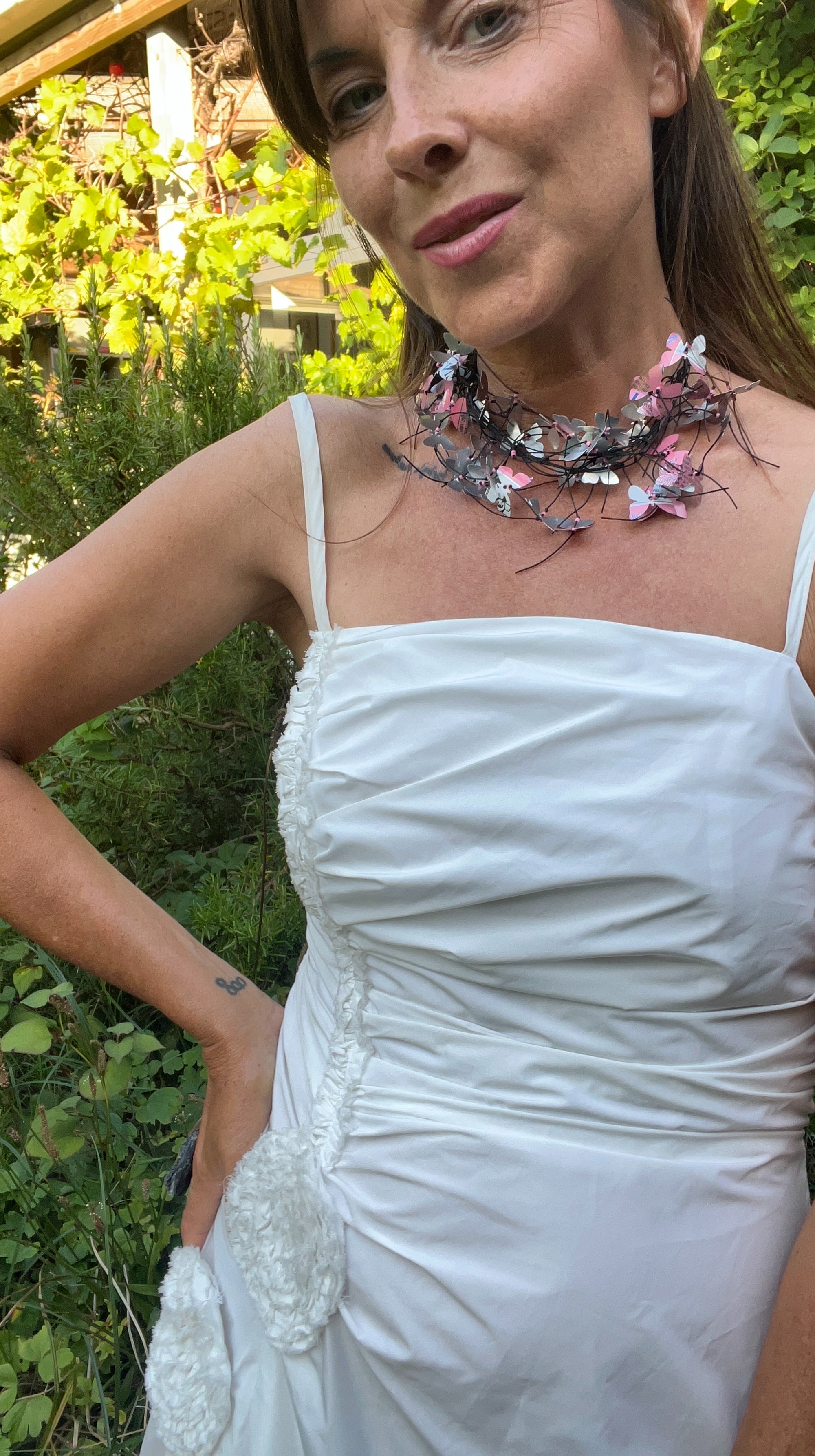 A woman outdoors wearing a white strap dress and a layered necklace made pink butterflies each one cut from soda cans, with green garden foliage in the background.