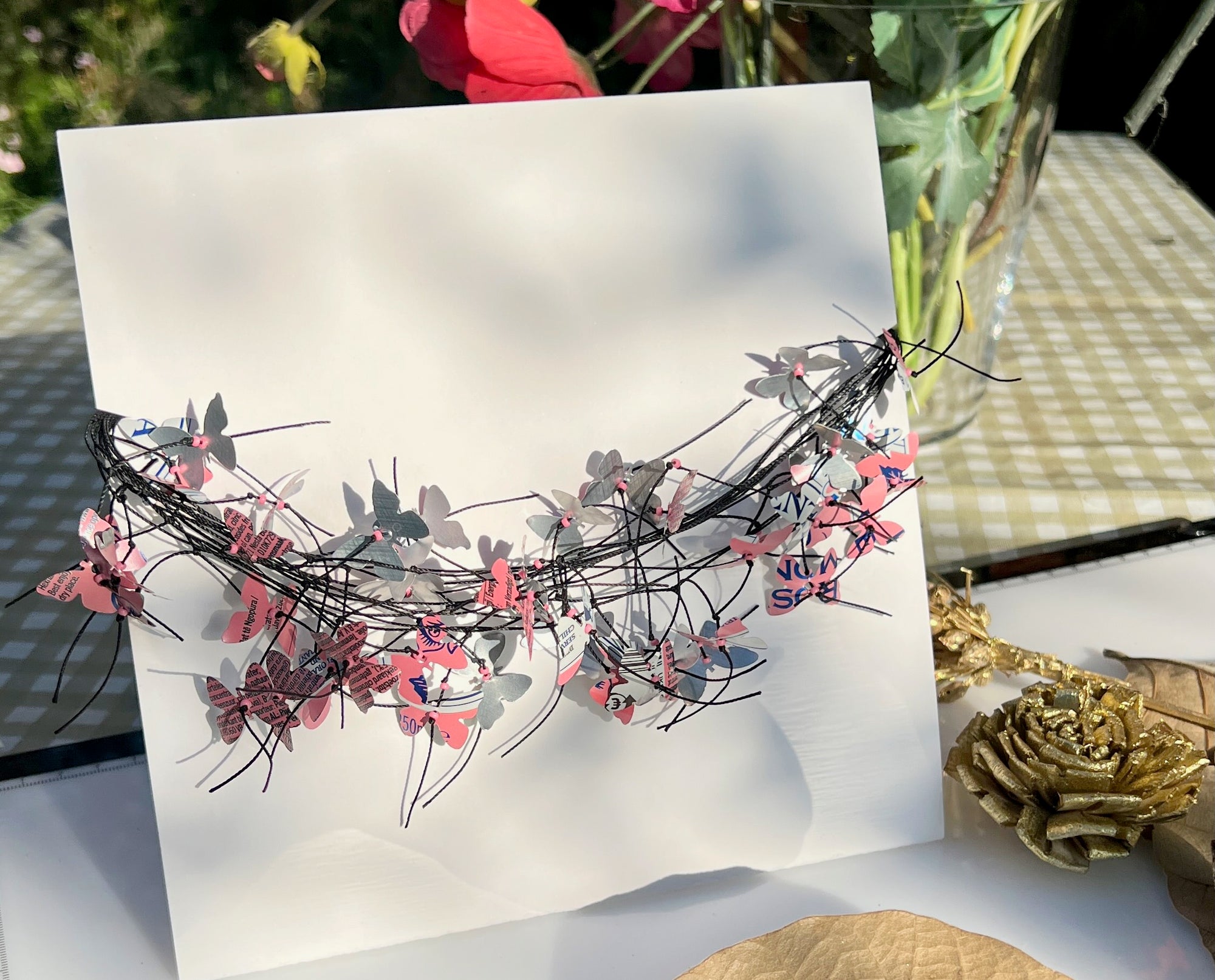 A handmade cord necklace decorated with small colorful aluminium butterflies cut from soda cans, displayed on a white card in a styled setting with flowers and a gold rose.