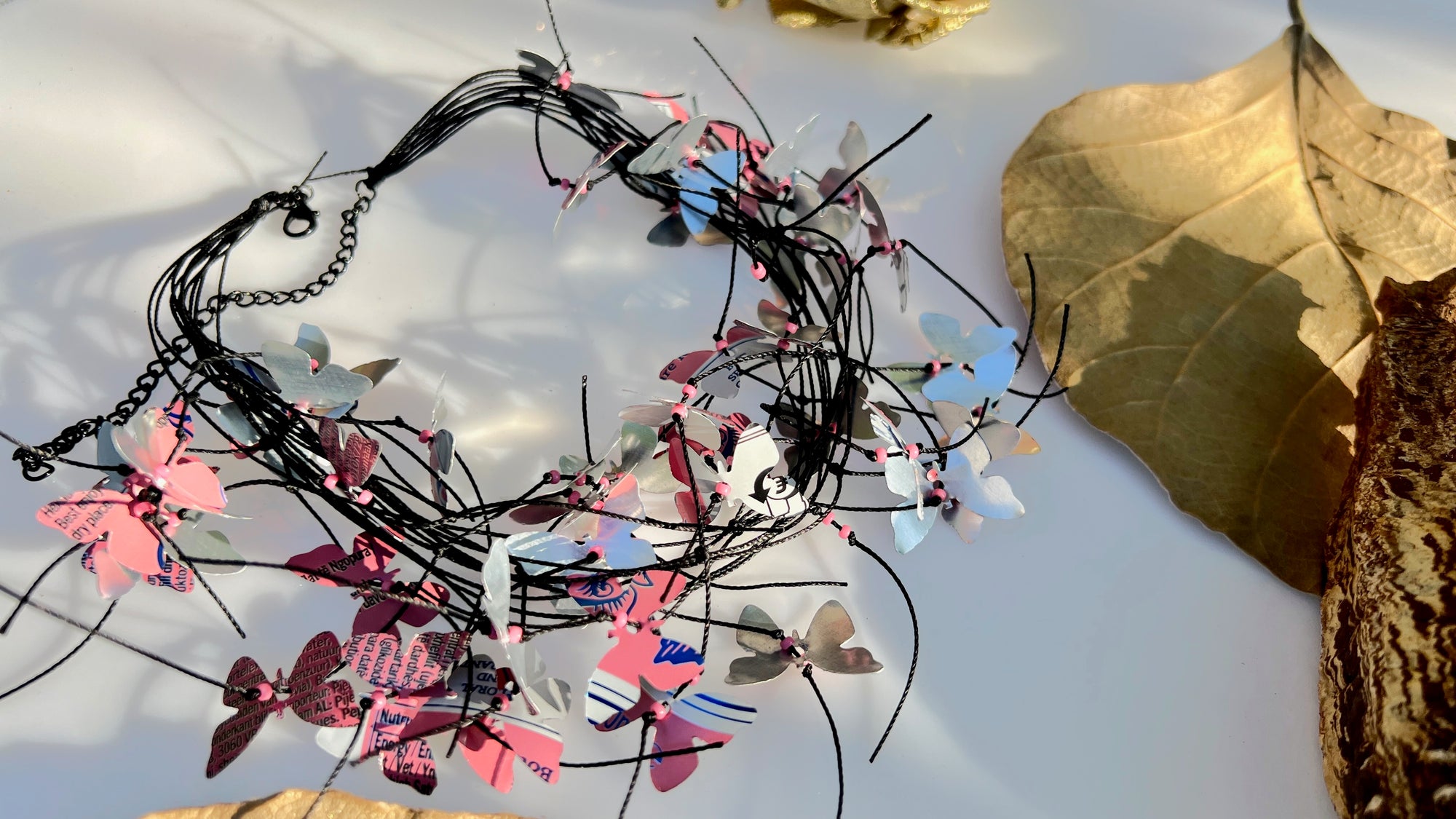 A handmade cord necklace decorated with small colorful aluminium butterflies cut from soda cans, arranged on a white surface with dried leaves.