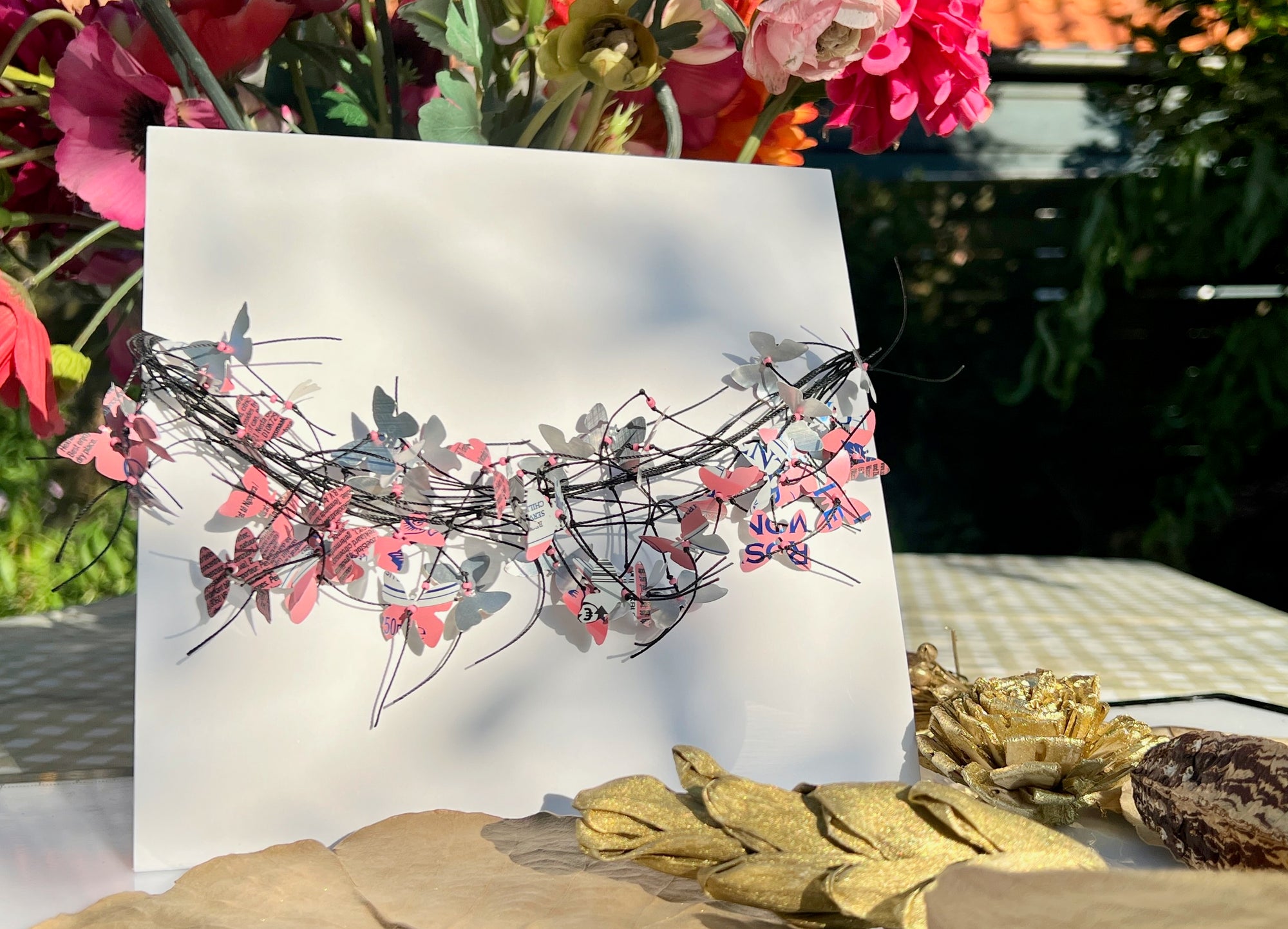 A white rectangular card or print featuringA handmade cord necklace decorated with small colorful aluminium butterflies cut from soda cans,  displayed outdoors on a table with flowers and dried leaves in the background.