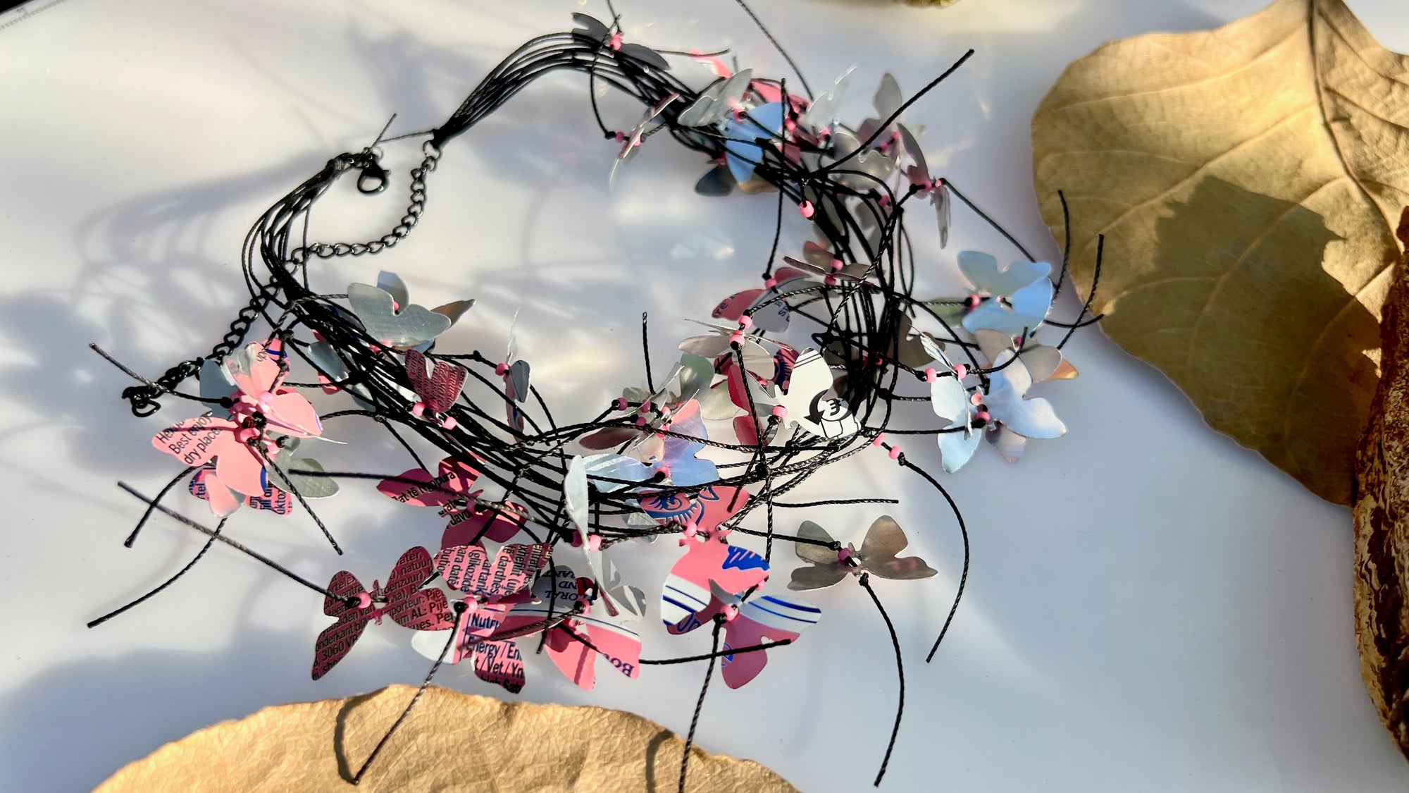 A handmade cord necklace decorated with small colorful aluminium butterflies cut from soda cans, styled on a white surface with dried leaves.