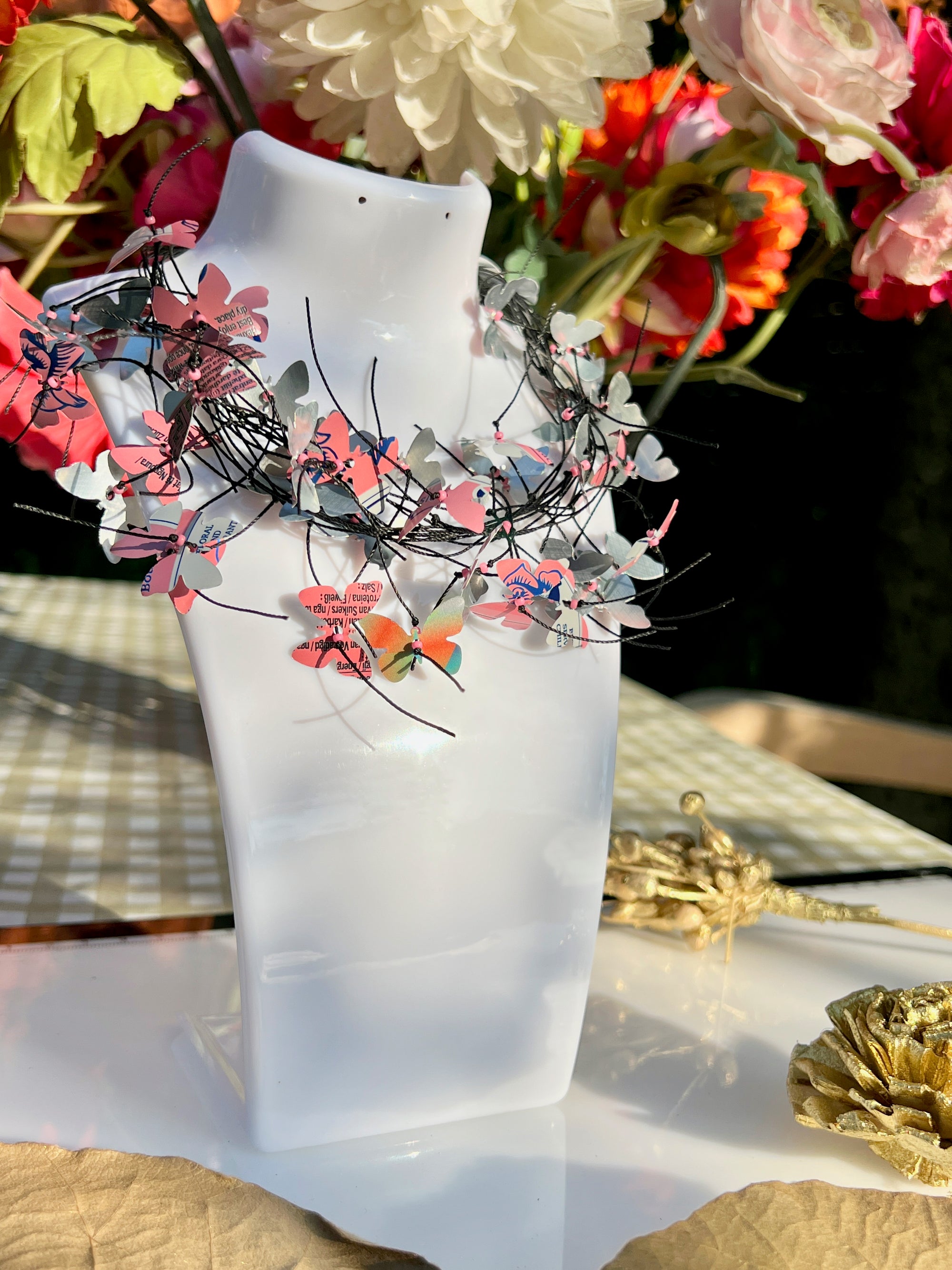 A white mannequin bust displays A handmade cord necklace decorated with small colorful aluminium butterflies cut from soda cans, set against a background of colorful artificial flowers and a checkered tablecloth.