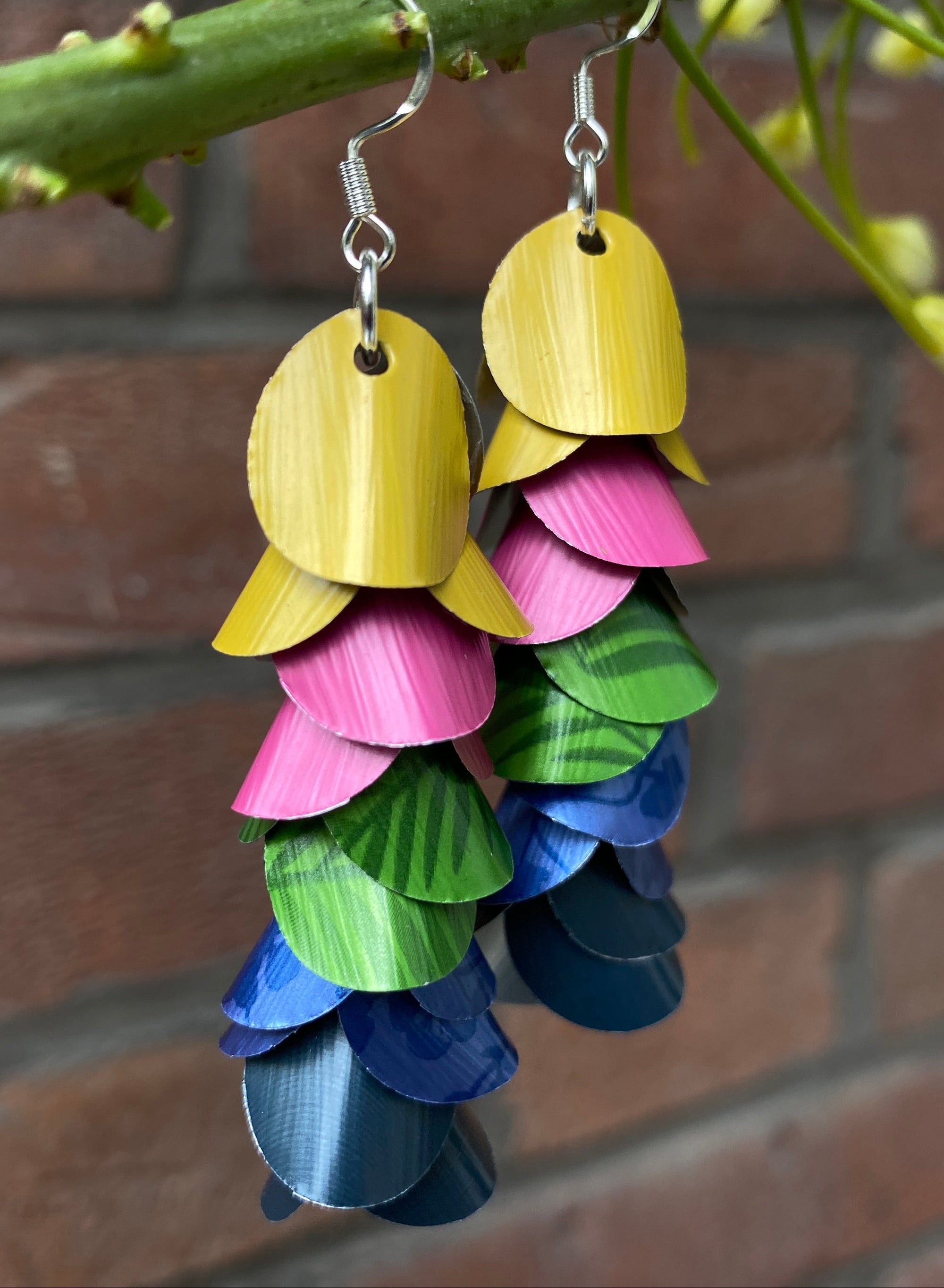 Colorful layered earrings made from upcycled nespresso capsules hanging from a branch with a brick wall background