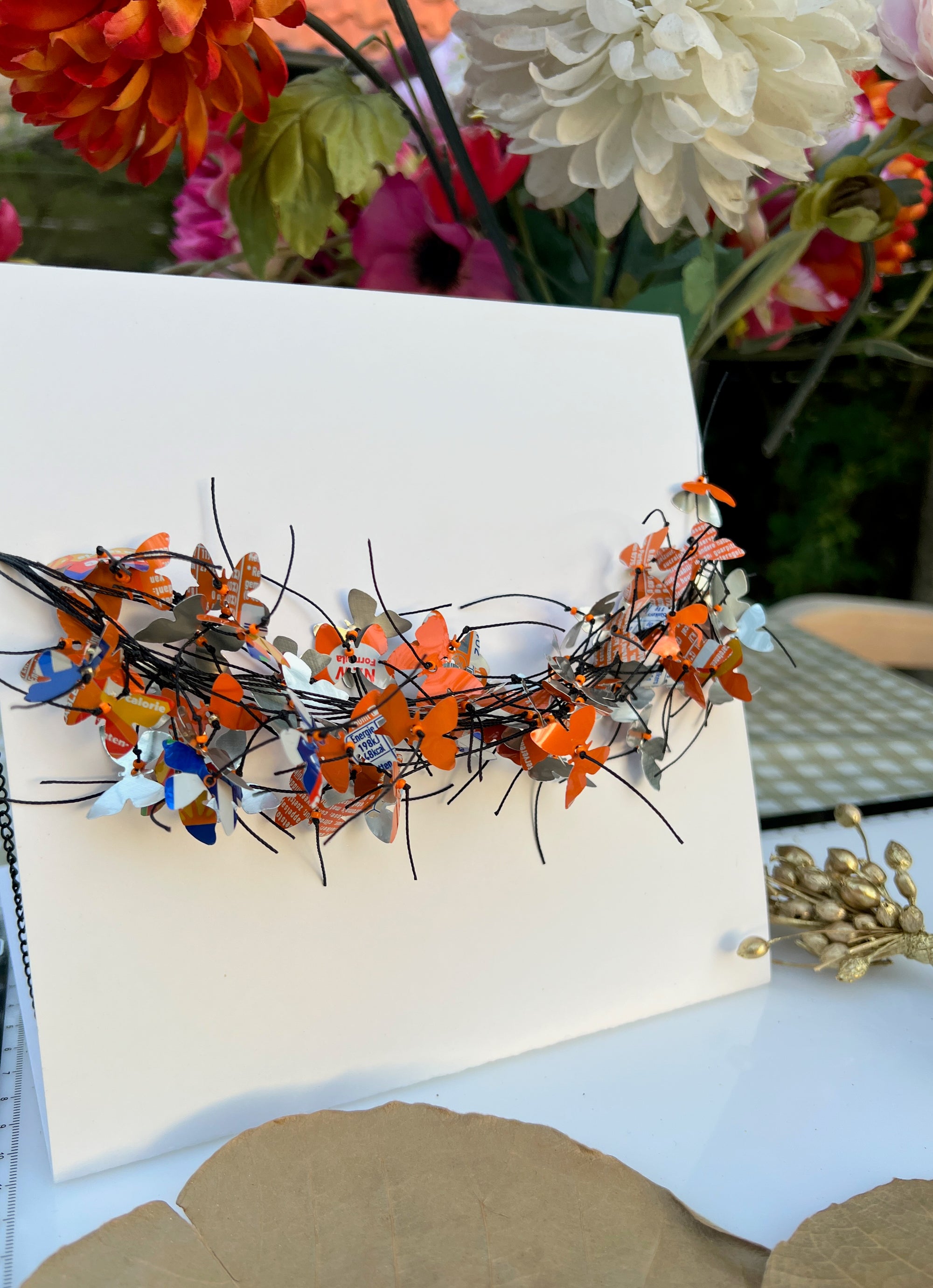 A white square card or board displayed on a table, featuring a decorative arrangement of orange, blue butterfly-shaped necklace attached across the center. The necklace is made from recycled fanta cans. The background shows blurred flowers and tabletop decor.