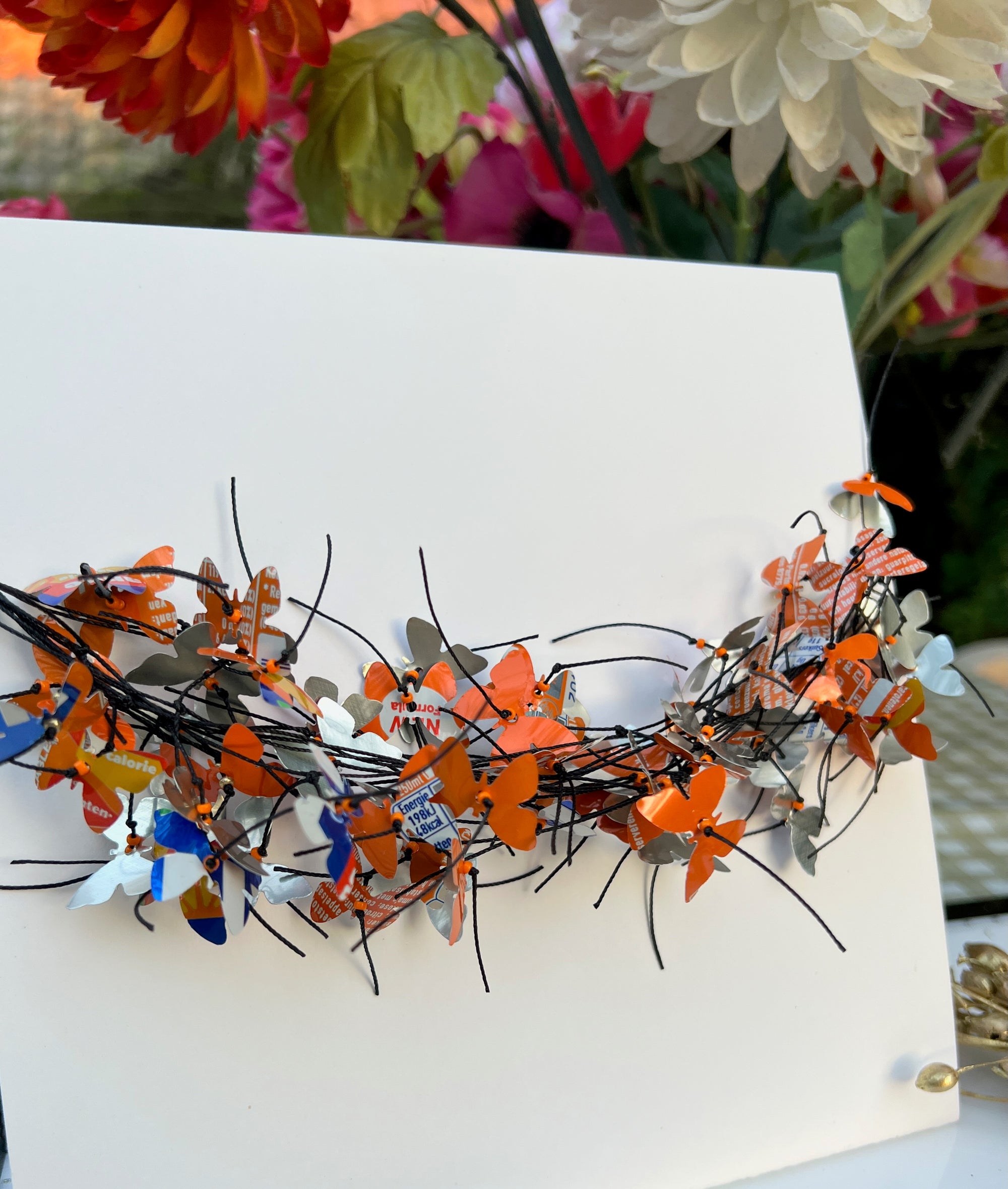 A white square card or board displayed on a table, featuring a decorative arrangement of orange, blue butterfly-shaped necklace attached across the center. The necklace is made from recycled fanta cans. The background shows blurred flowers and tabletop decor.