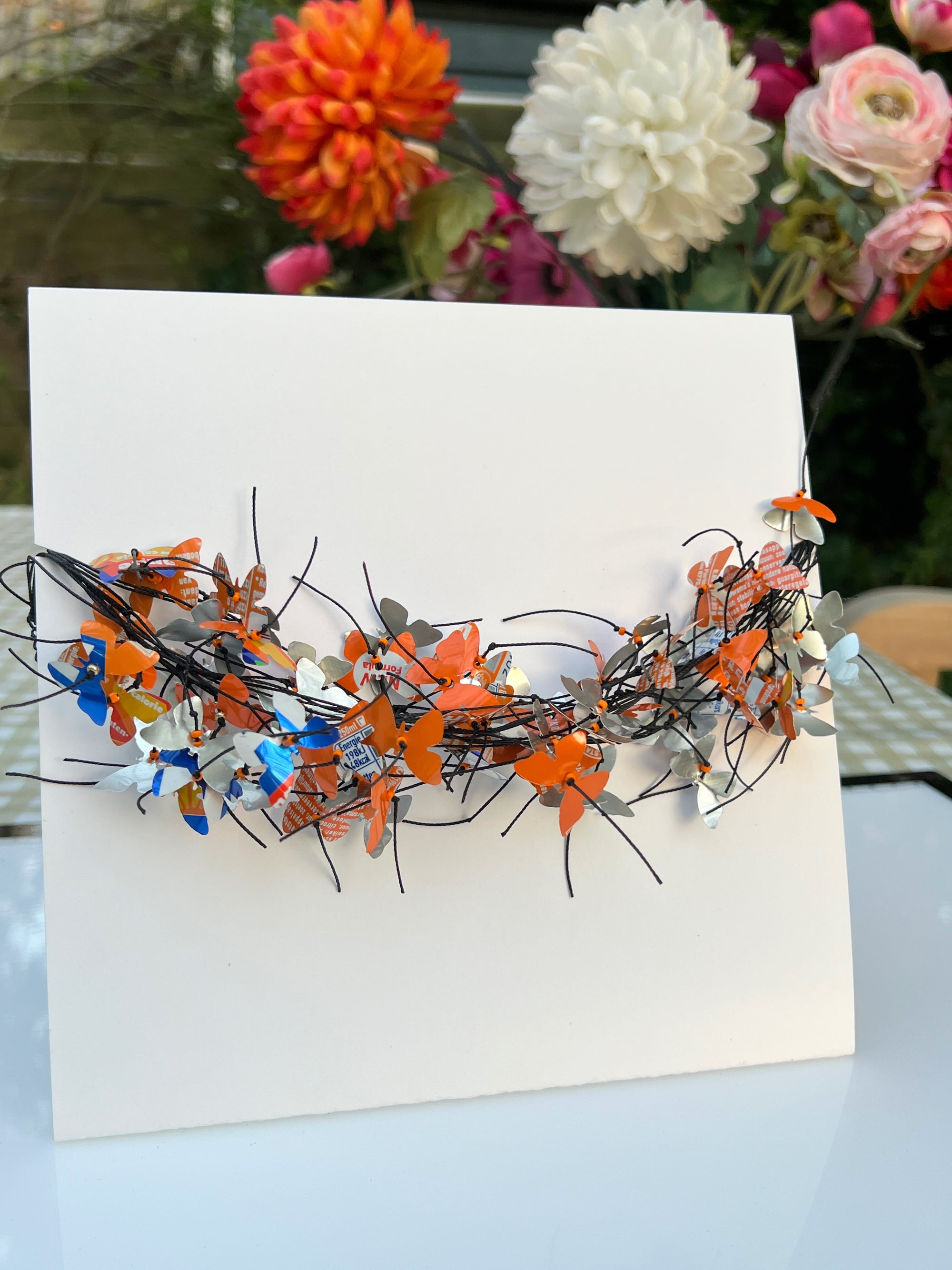 A white square card or board displayed on a table, featuring a decorative arrangement of orange, blue butterfly-shaped necklace attached across the center. The necklace is made from recycled fanta cans. The background shows blurred flowers