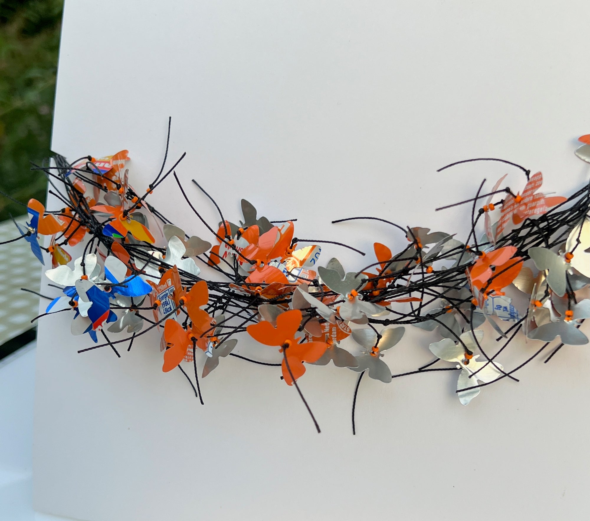 A white square card or board displayed on a table, featuring a decorative arrangement of orange, blue butterfly-shaped necklace attached across the center. The necklace is made from recycled fanta cans. 