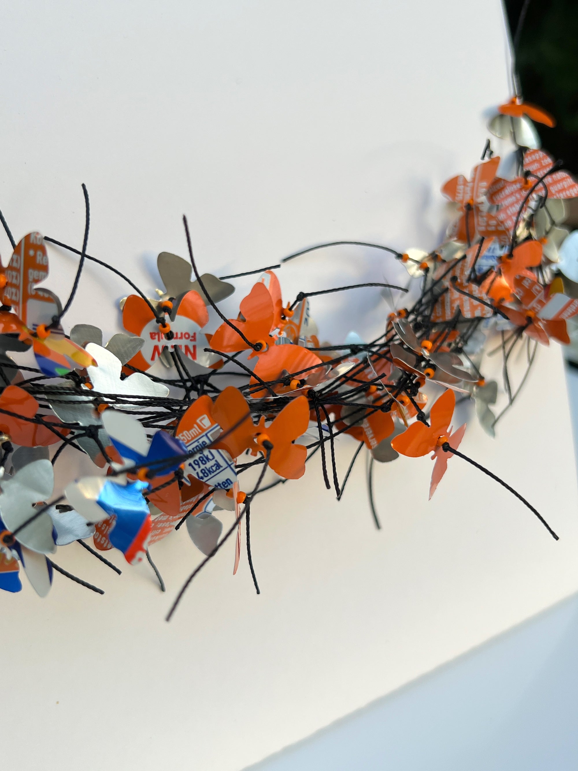 A close-up of a decorative necklace made of many small butterfly-shaped pieces in orange, silver, white, blue, and red, attached to thin black cord, photographed against a plain white background. The necklace is made from Fanta cans