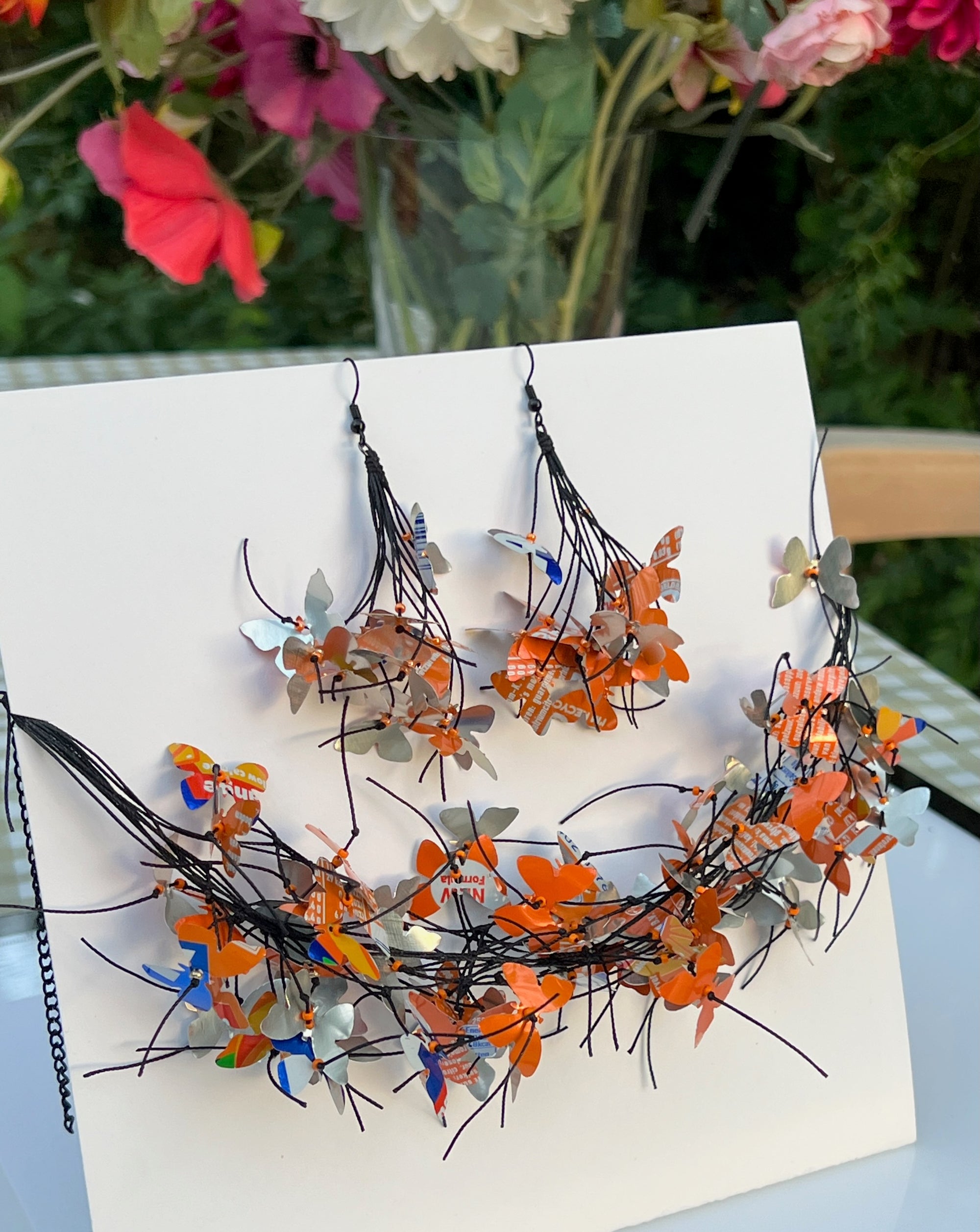 A handmade jewelry set featuring a crescent-shaped wire necklace and matching drop earrings adorned with numerous small orange, silver, and blue butterfly cutouts, displayed on a white card. The jewelry is made from repurposed Fanta cans 
