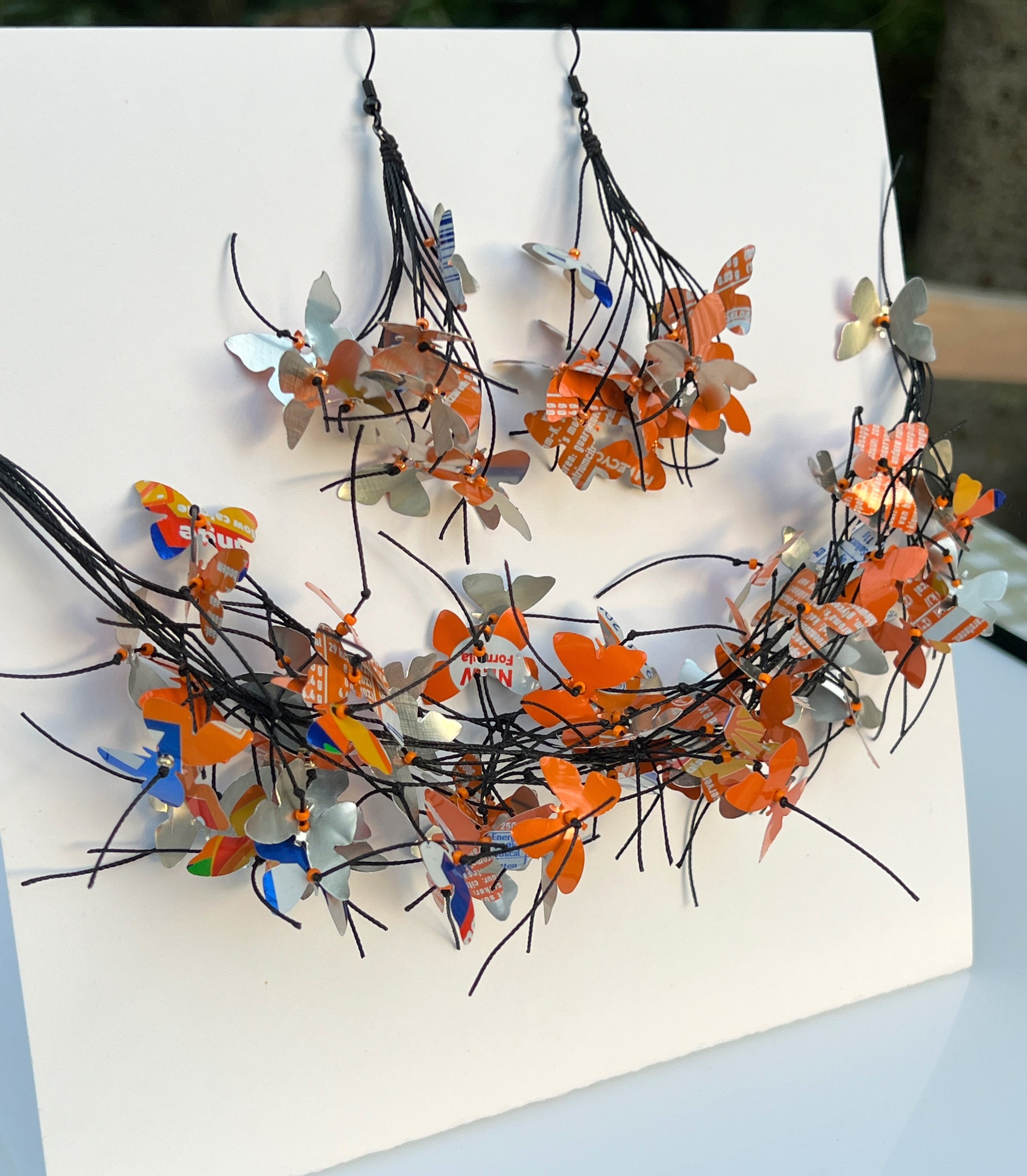 A handmade butterfly garland and matching earrings made from colorful recycled paper and metallic foil butterflies attached to black string, displayed on a white card. The jewelry is made from recycled fanta cans. 