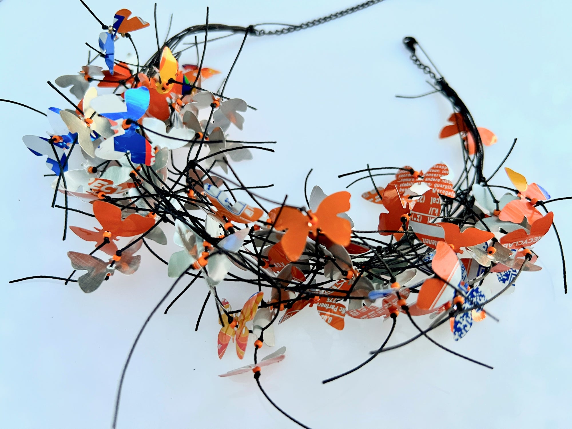 A handmade butterfly crown or headpiece featuring numerous colorful aluminum butterflies cut from fanta cans, in orange, blue, and silver attached to a black wire headband, displayed on a light blue background.