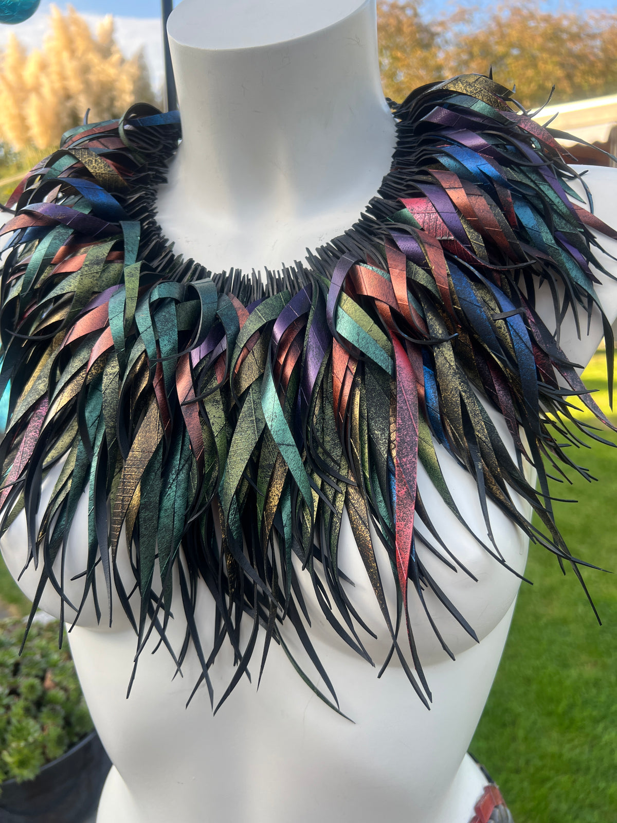 A large, statement collar necklace made of layered, iridescent rubber strips in shades of teal, green, blue, purple, pink, and black, displayed on a white mannequin torso outdoors. The necklace is made of upcycled bicycle innertube rubber. 