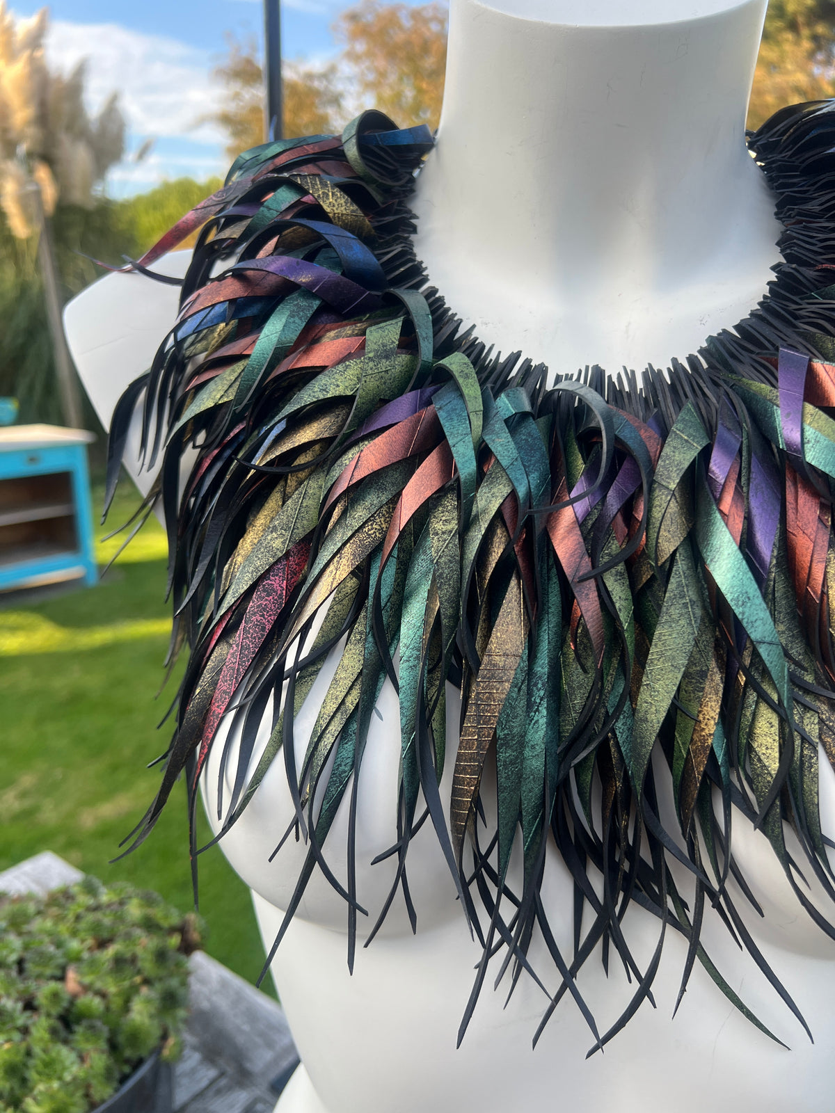 A large statement collar necklace made of layered iridescent feather-shaped strips, displayed on a white mannequin outdoors with a blurred garden background.