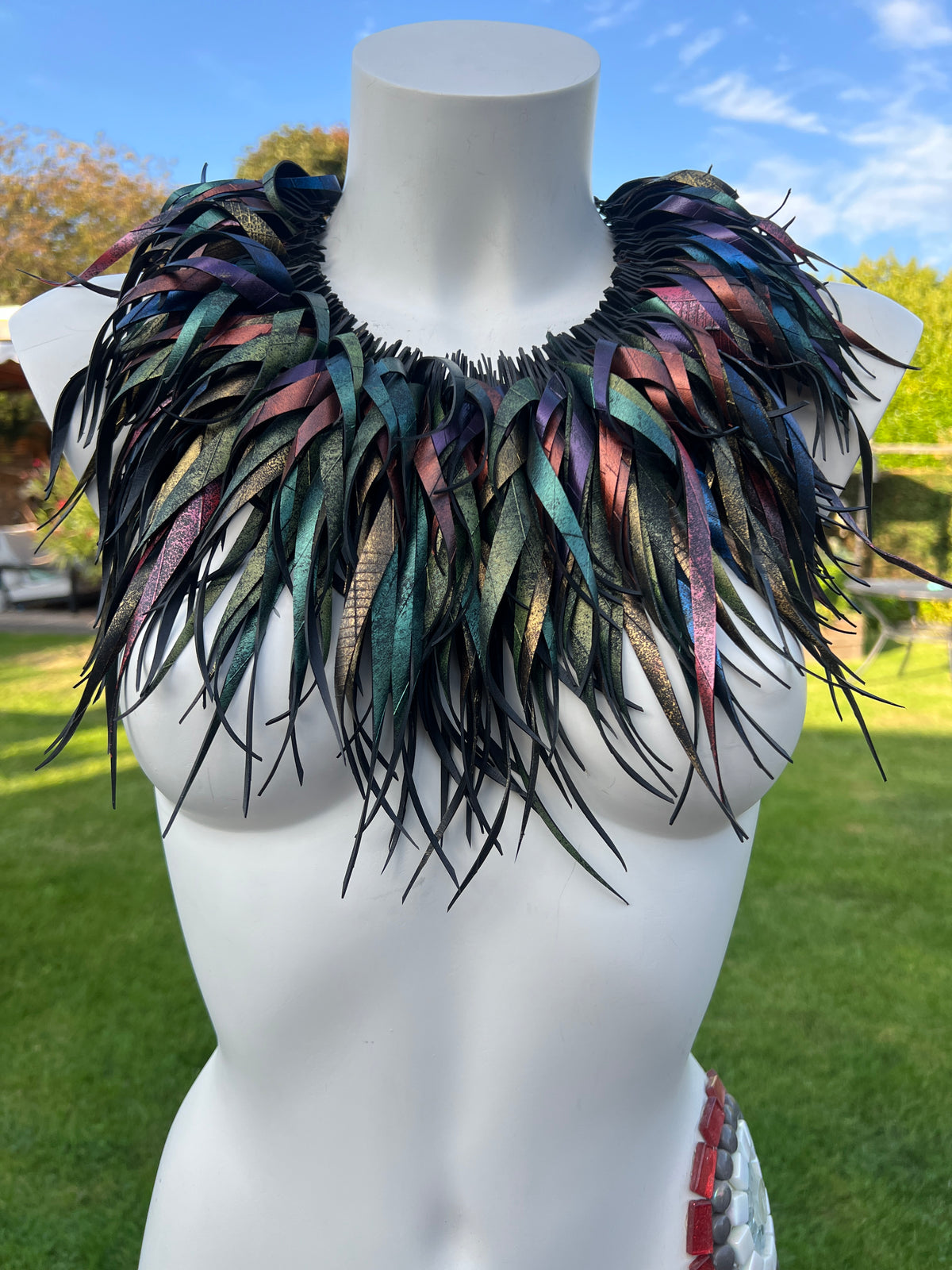 A large, dramatic feathered collar necklace displayed on a white mannequin torso outdoors. The collar features layered curly tassels, made from repurposed bicycle inner tube rubber in black with iridescent, metallic hues of green, blue, purple, and pink.