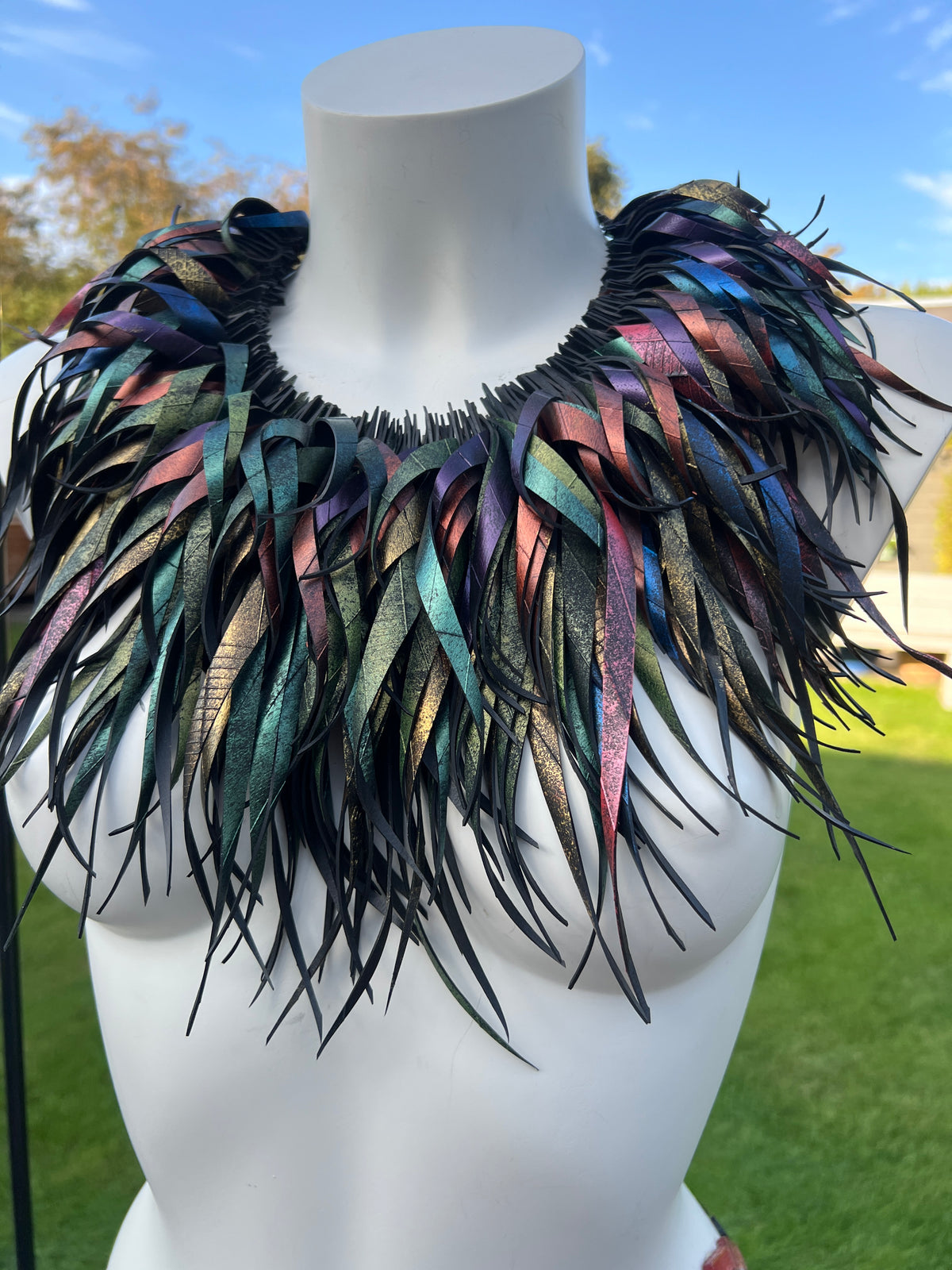 A large statement collar necklace made of layered iridescent upcycled bike inner tube rubber strips, displayed on a white mannequin outdoors with grass and blue sky in the background.
