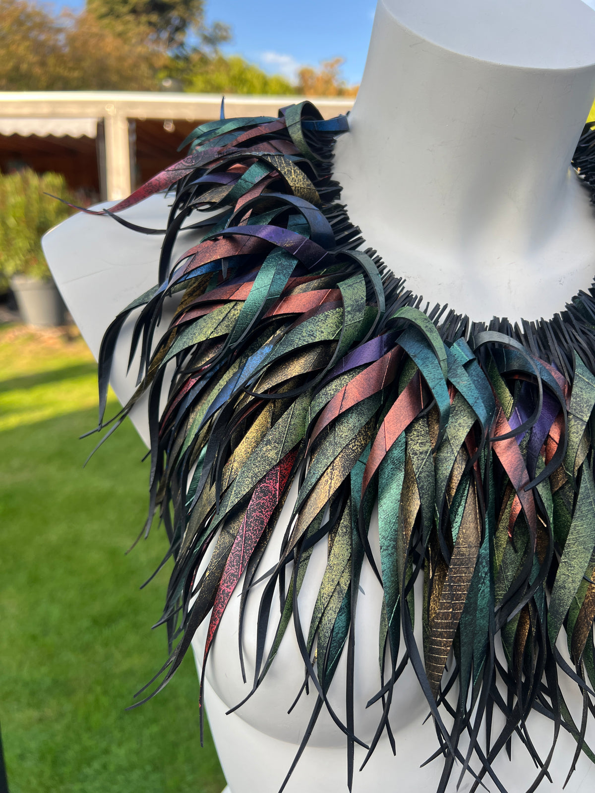 A large statement necklace made of layered, iridescent dark curly strips, made from repurposed bike inner tube rubber, displayed on a white mannequin torso outdoors with a green lawn and house in the background.