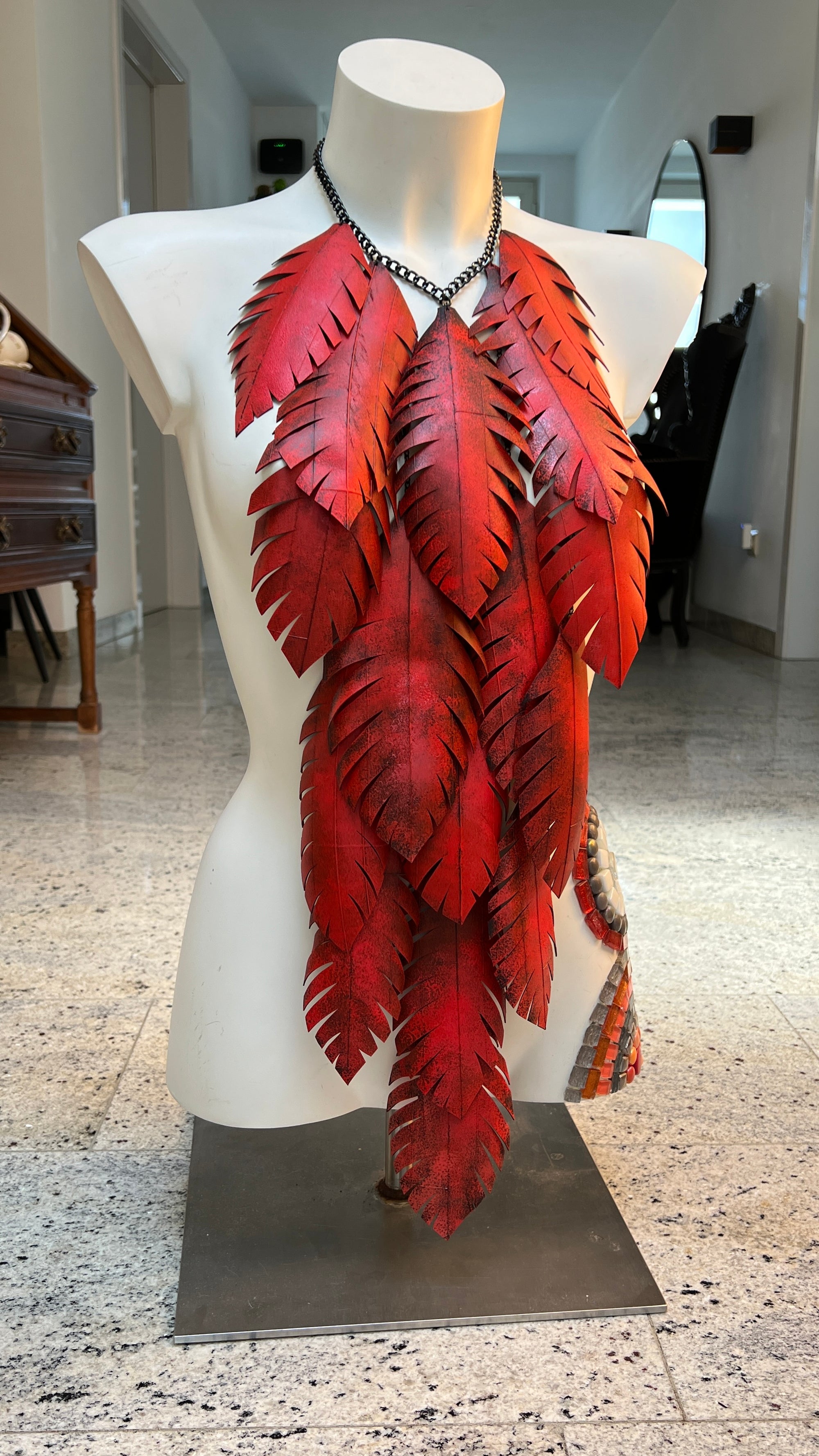 very large, red coloured, layered leaf shaped statement necklace made from upcycled bike tyre rubber on a white mannequin in a home environment